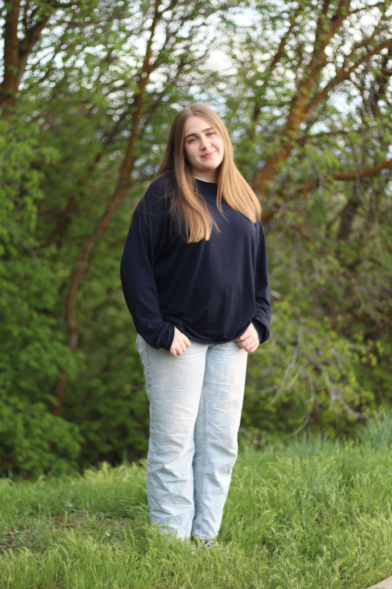 Teen girl with long hair wearing navy sweatshirt and gray jeans standing in green nature setting.