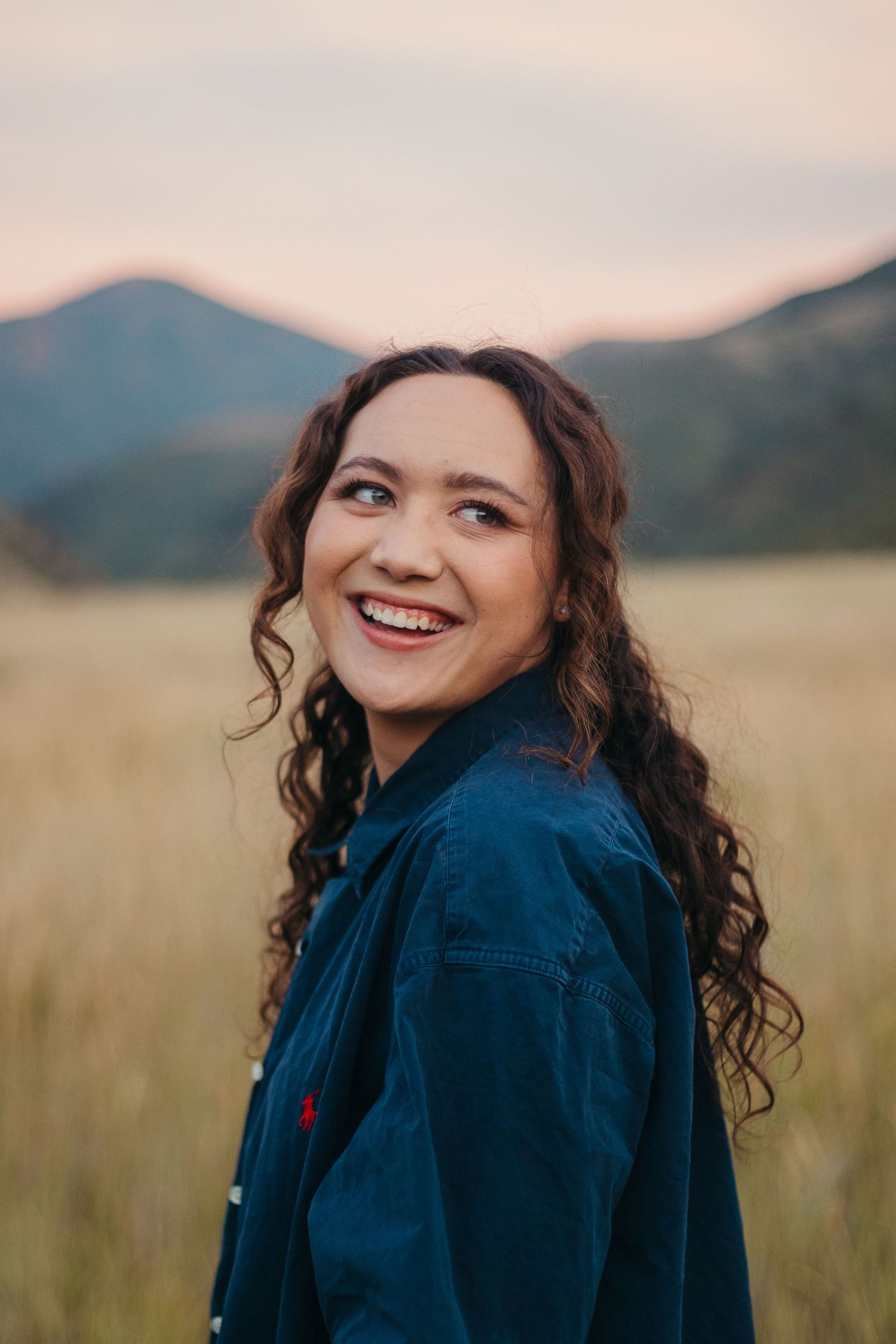 Young woman with curly hair in dark blue jacket smiling over shoulder in golden field at sunset.