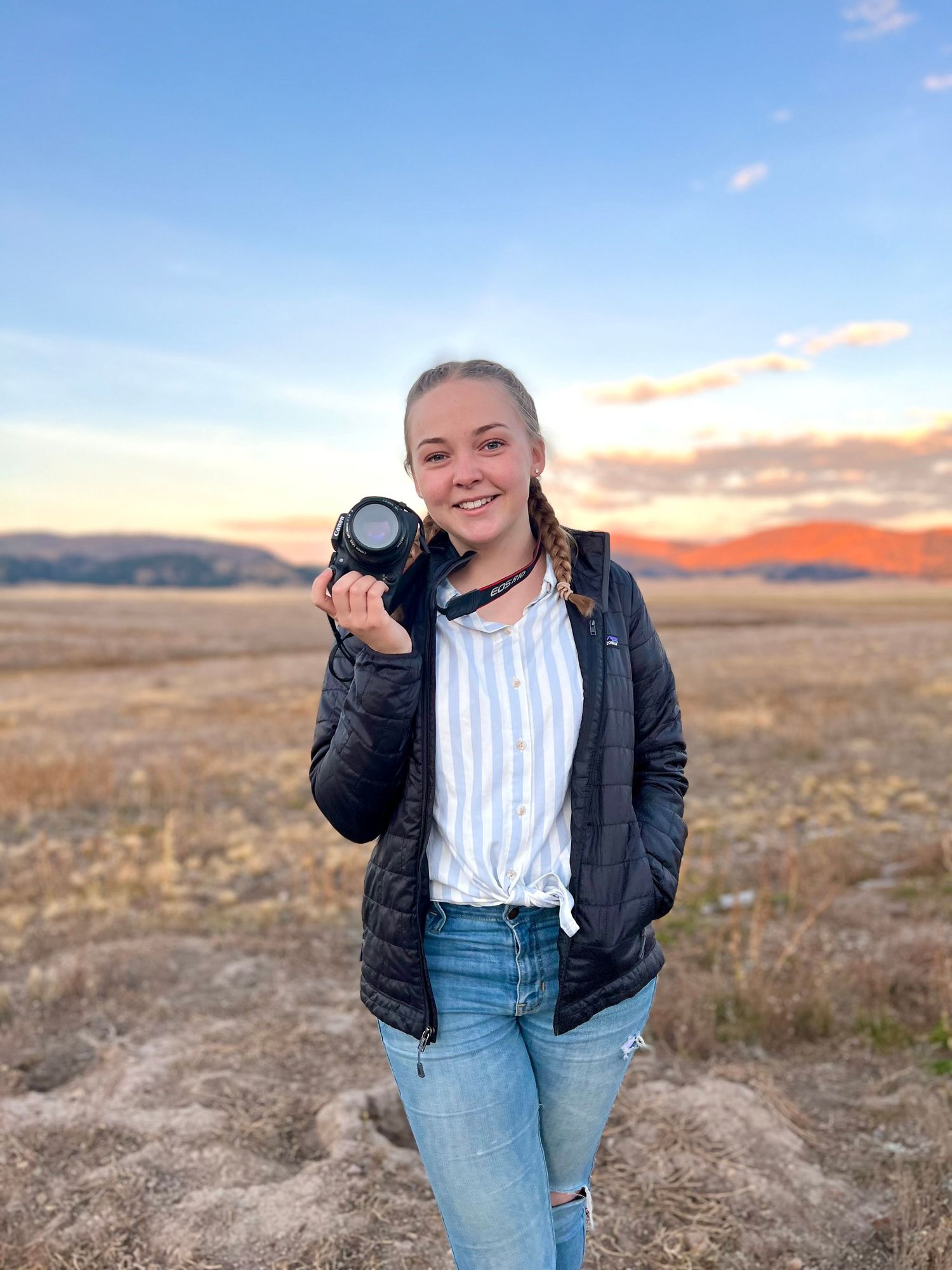 Young girl holding a camera lens smiling in an open field at sunset with mountains in the background.