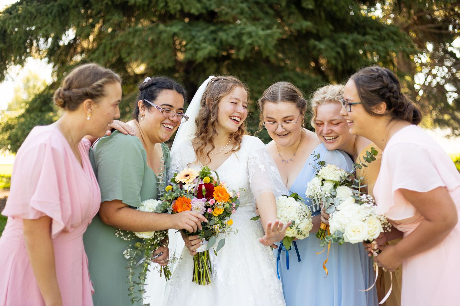 Bride laughing with bridesmaids in colorful dresses holding bouquets outdoors at a wedding celebration.