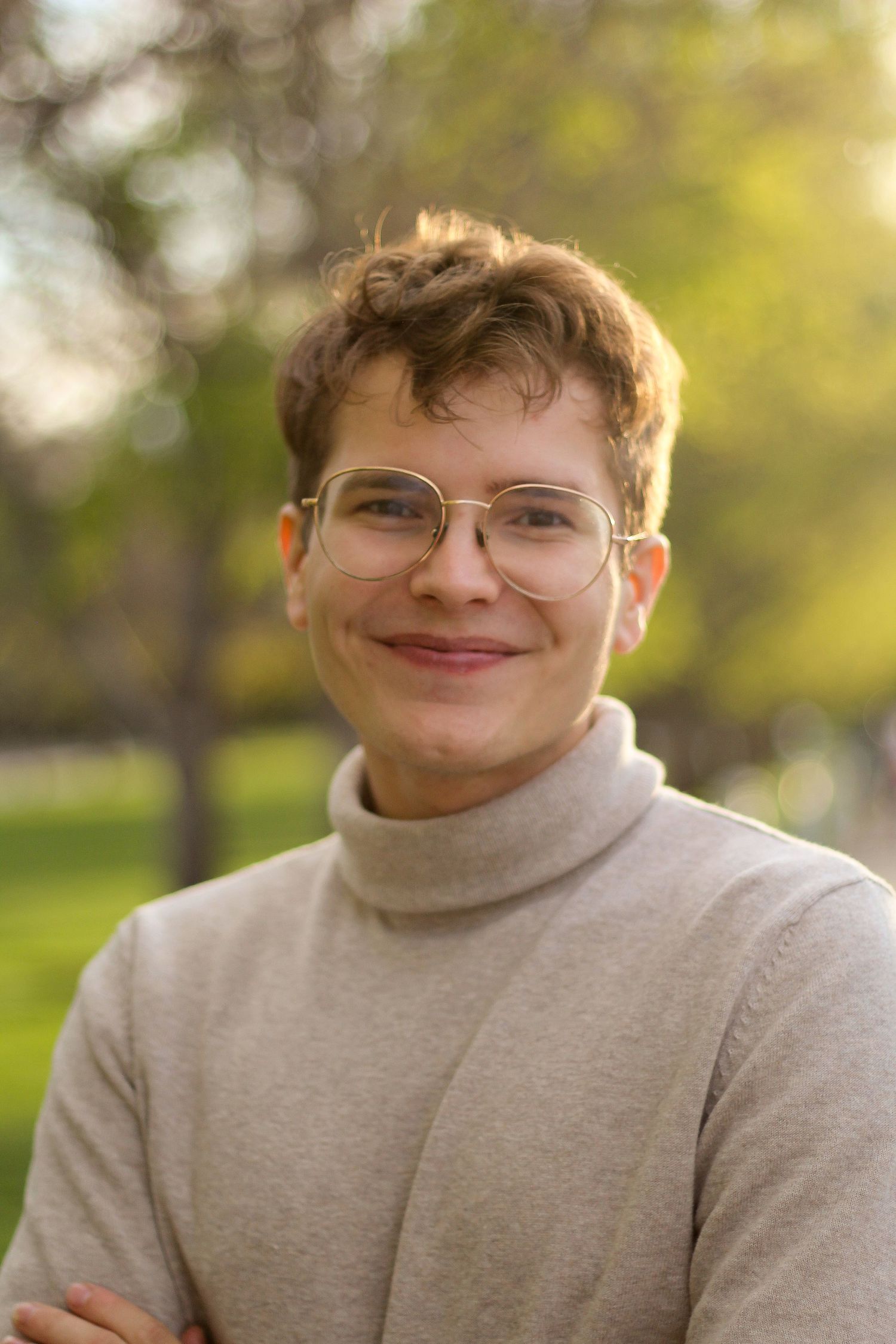 Young person with short auburn hair and glasses wearing a beige turtleneck sweater smiling outdoors in a park.