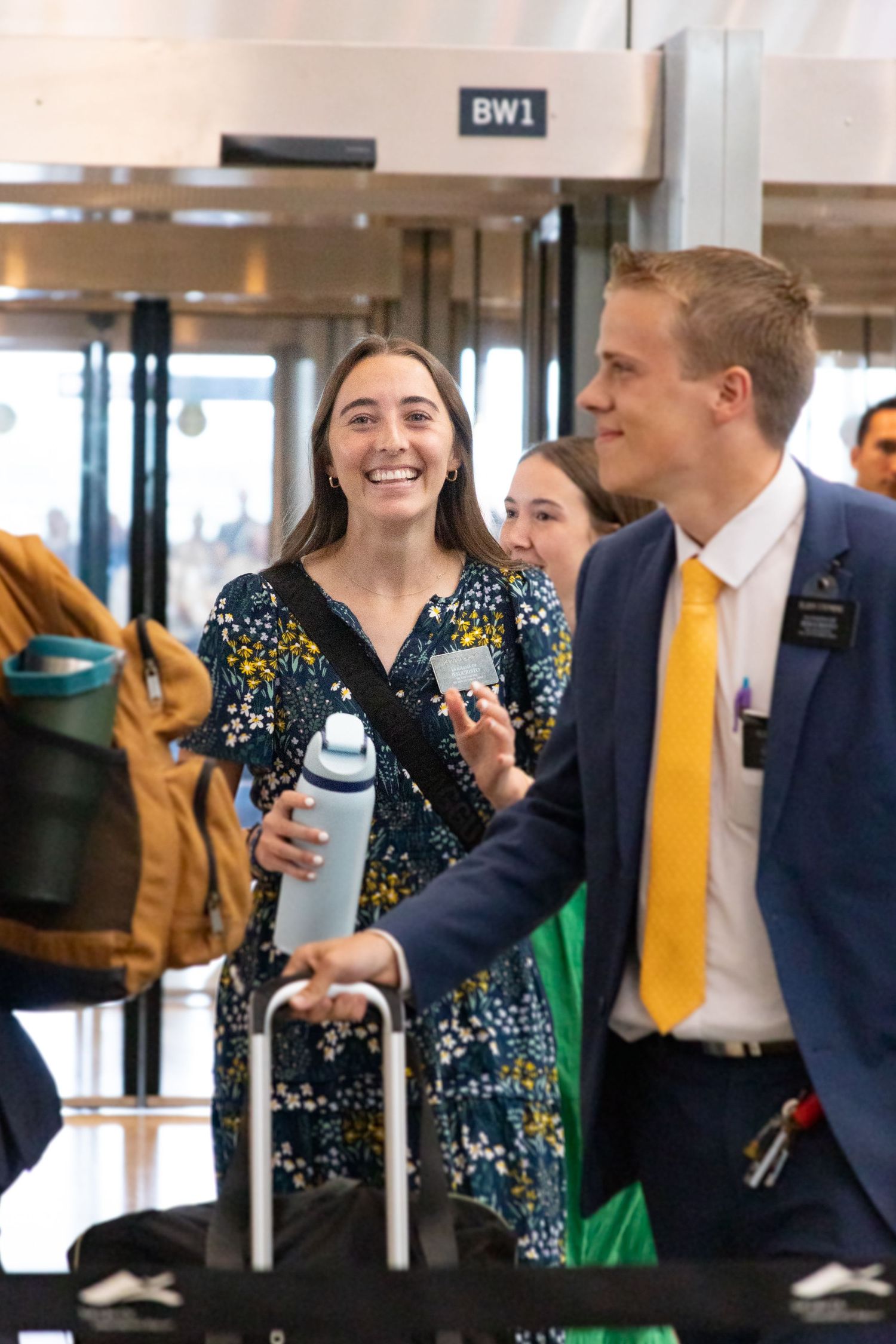 Smiling return missionary in floral dress walks through airport terminal with luggage.
