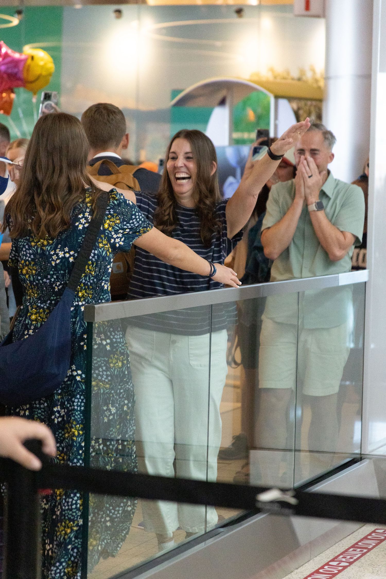 Emotional woman laughs and reaches over a glass barrier to greet her returned missionary daughter at an airport.