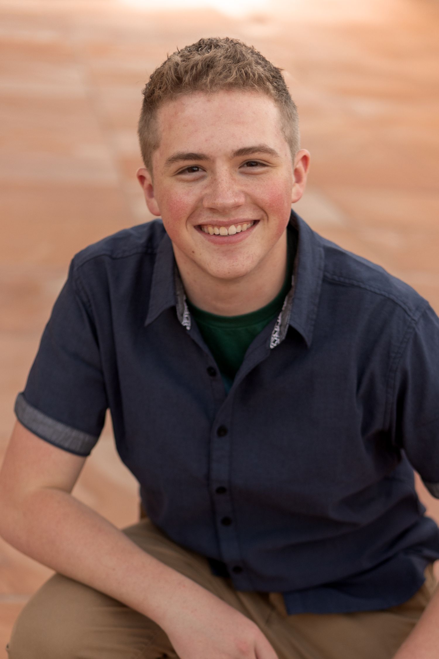 Smiling teenage boy with short hair wearing a navy blue button-up shirt, sitting outdoors against a warm brick background.