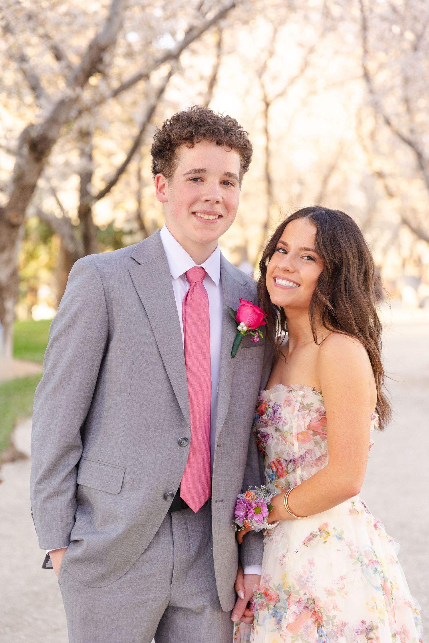 Prom couple posing among cherry blossoms, boy in gray suit with pink tie and girl in floral strapless dress.