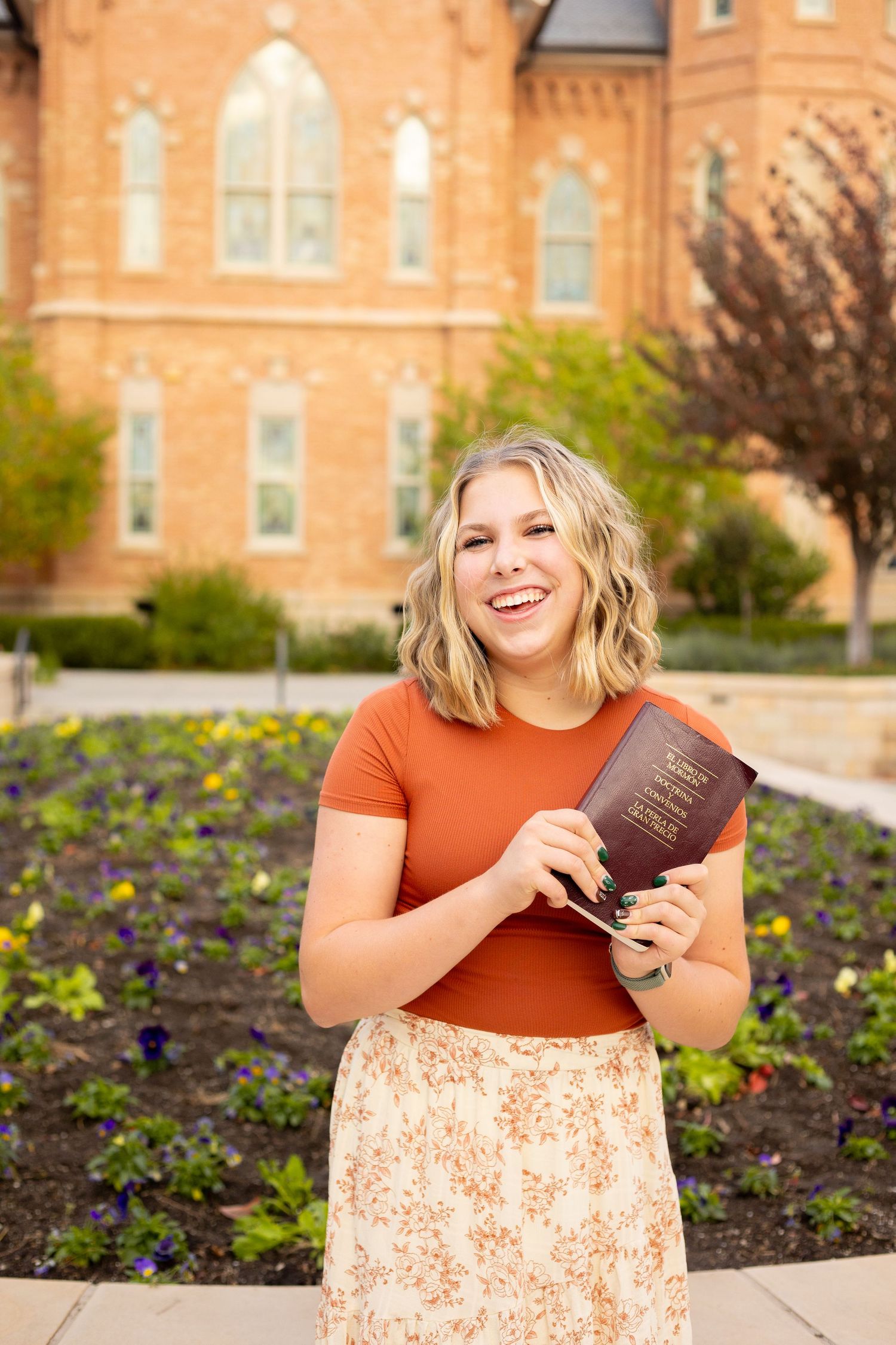 Laughing young woman in rust top holding book standing in front of brick church building.