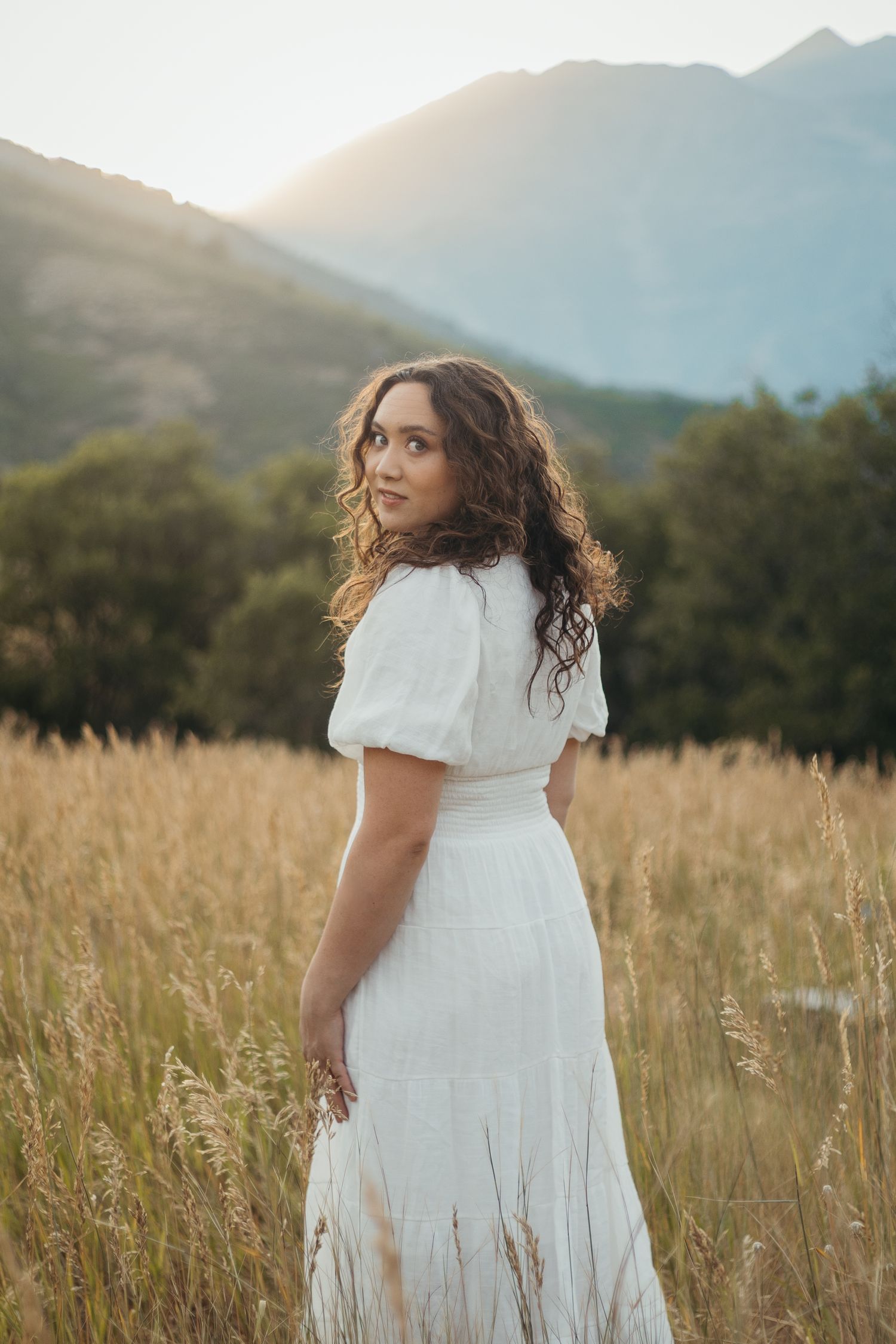 Woman in white puff-sleeve dress stands in golden wheat field with mountain sunset backdrop.