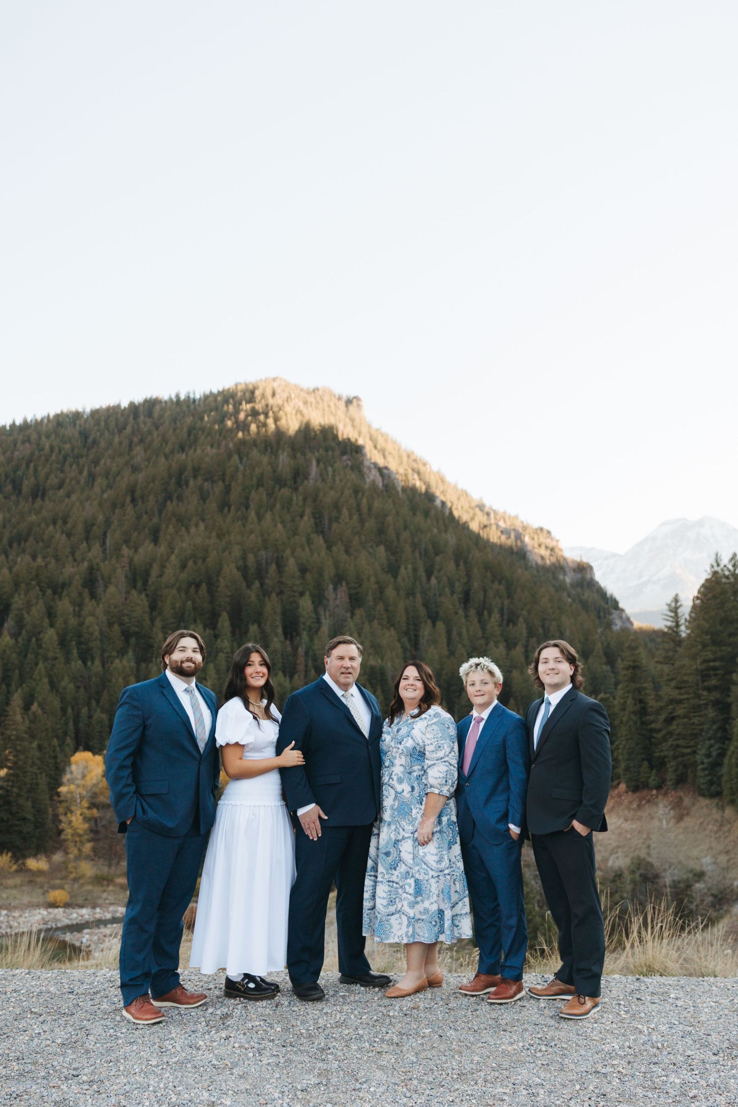 Six family members in formal attire posing outdoors with forested mountain at sunset.