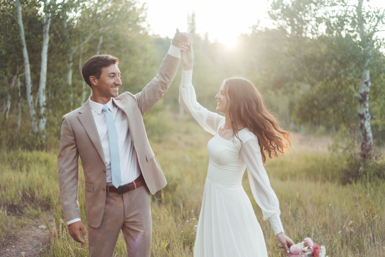 Groom spinning bride in white dress in a sunlit meadow surrounded by birch trees.