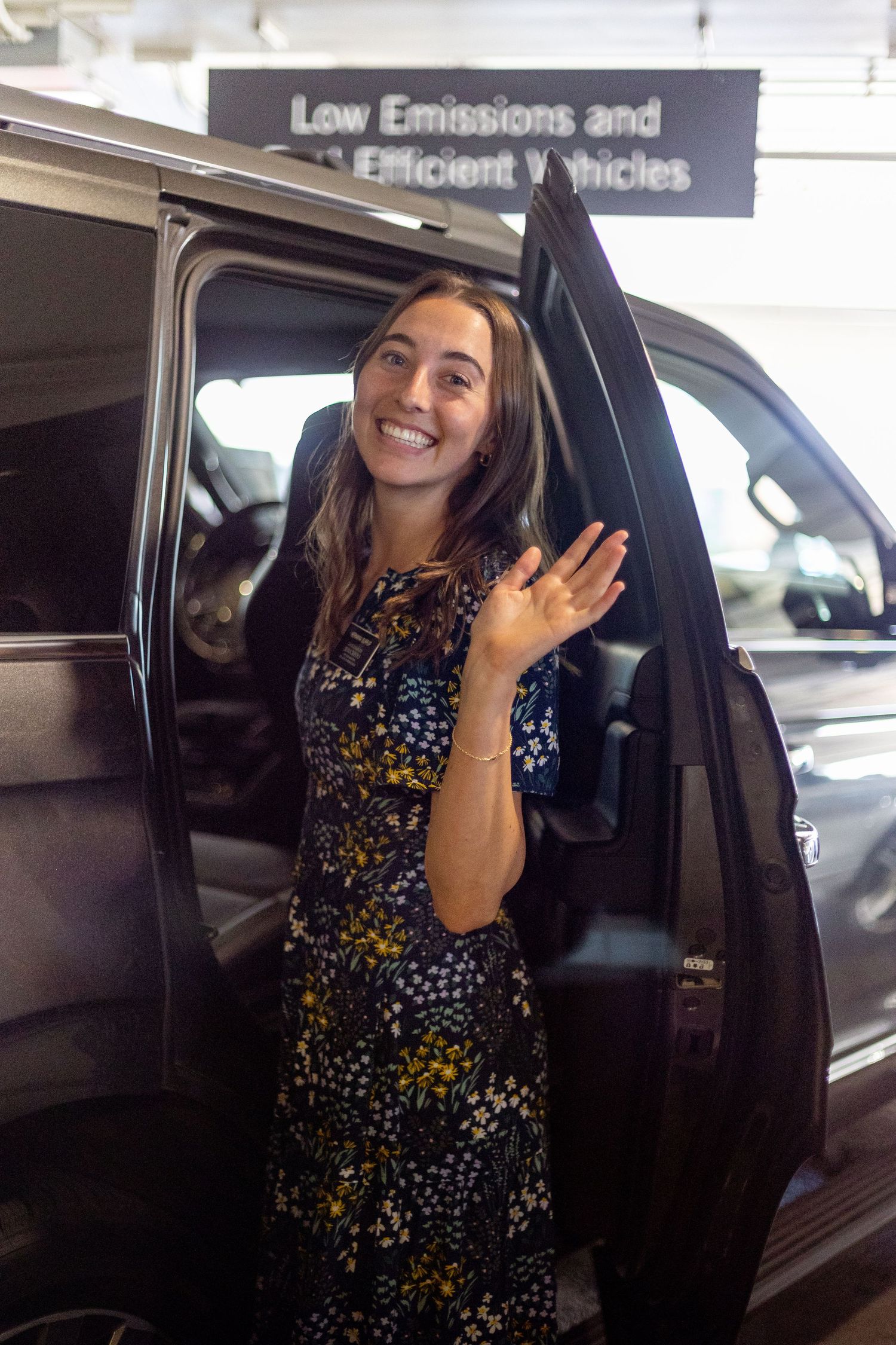 Smiling young woman in floral dress waving while standing beside a van in a parking garage.