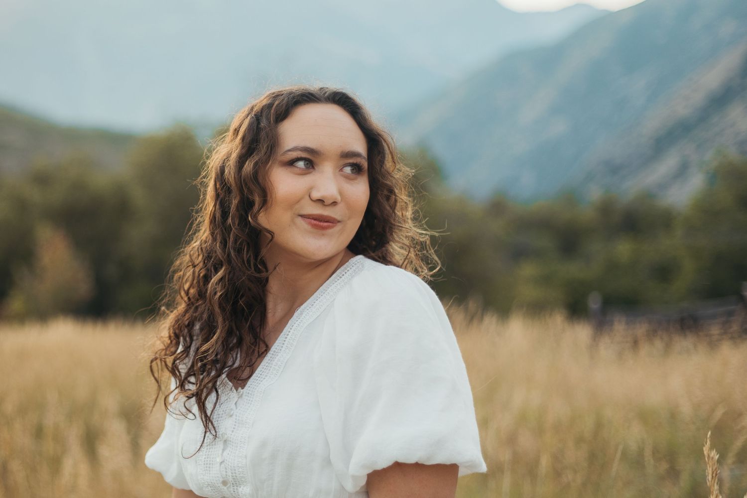Young woman with curly hair in white dress gazes over shoulder in golden meadow with mountain backdrop.