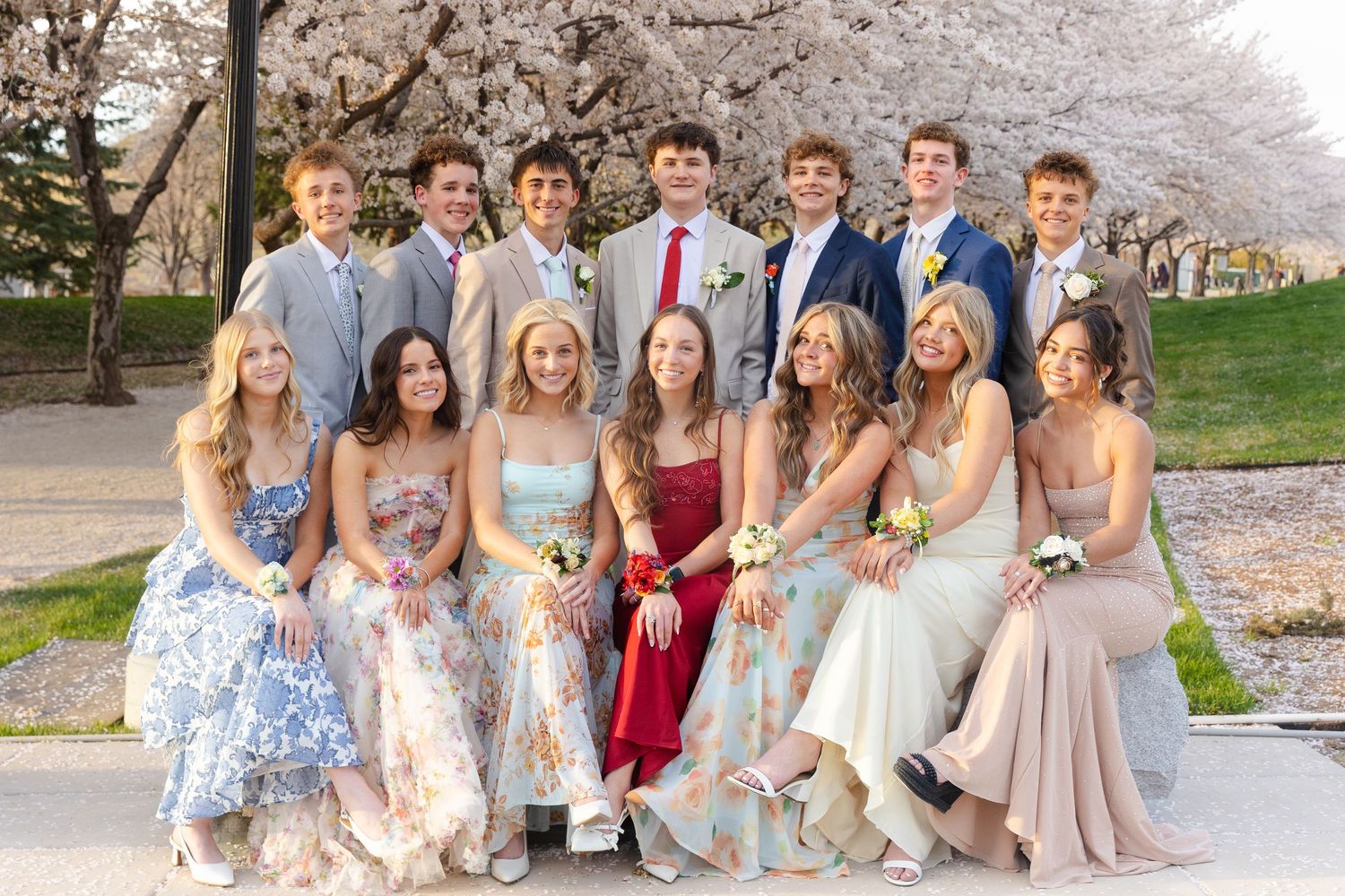 Smiling group of teenage prom couples posing together under white cherry blossom trees in formal wear at the Utah Capitol.