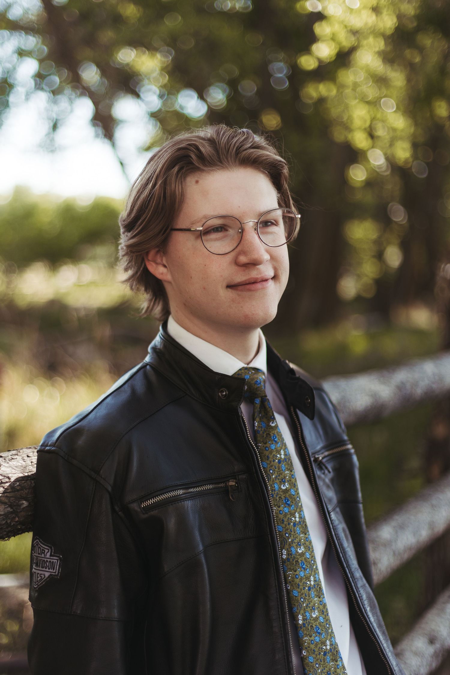 Young man in glasses, leather jacket, and floral tie smiling outdoors near a wooden fence.