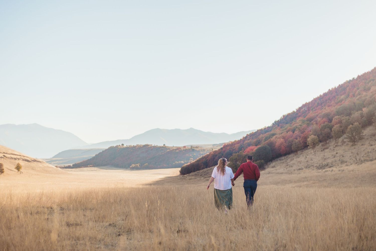 Couple walking hand in hand through golden meadow with colorful autumn mountains behind.