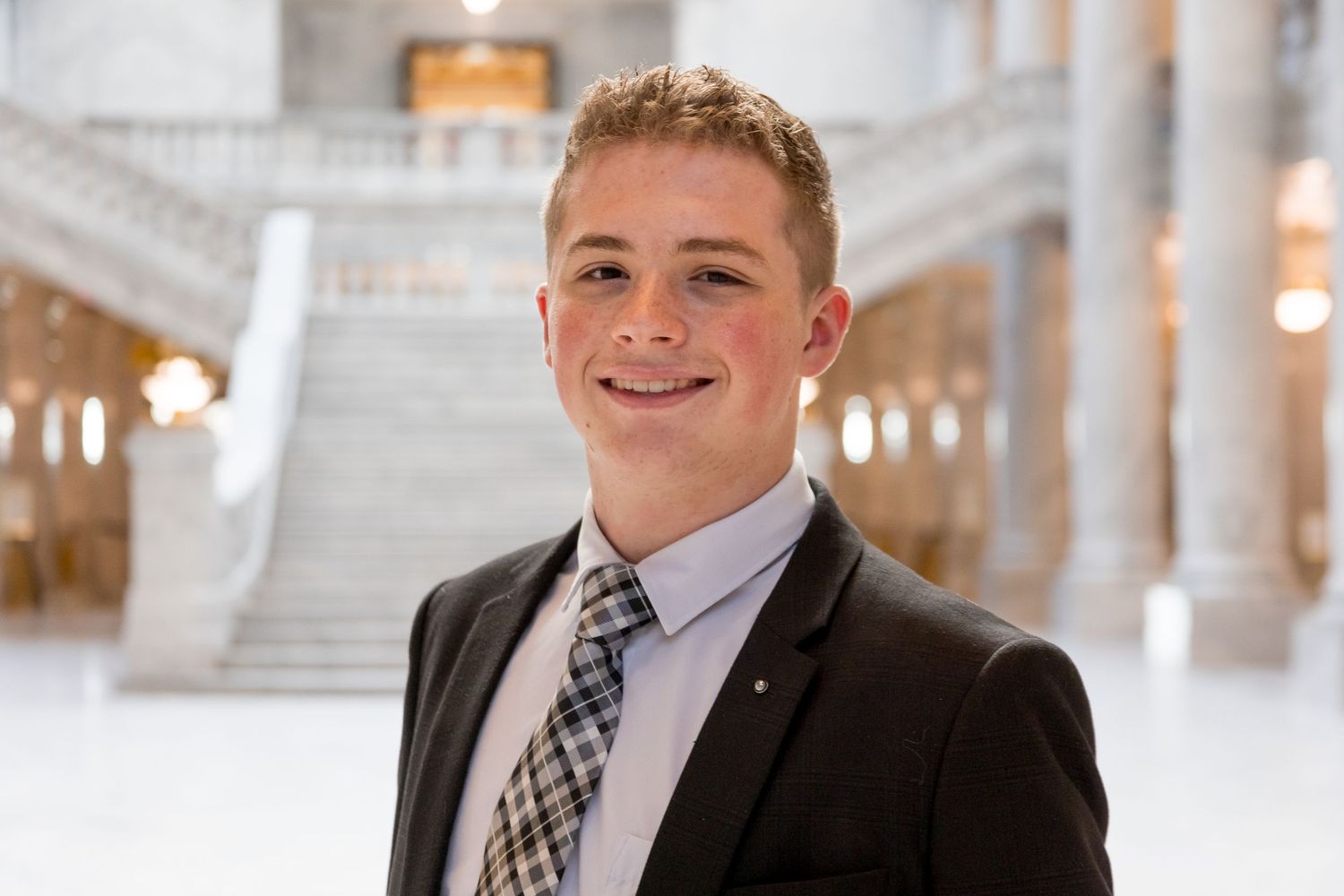 Smiling young man in a black suit and plaid tie posing inside a grand marble building interior.