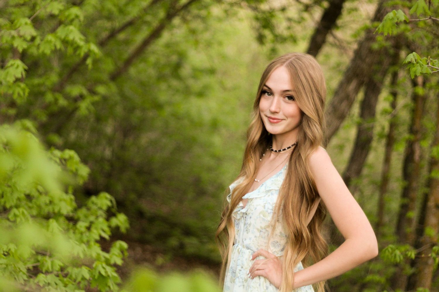Young woman with long blonde hair posing in a lush green forest during senior portrait session.