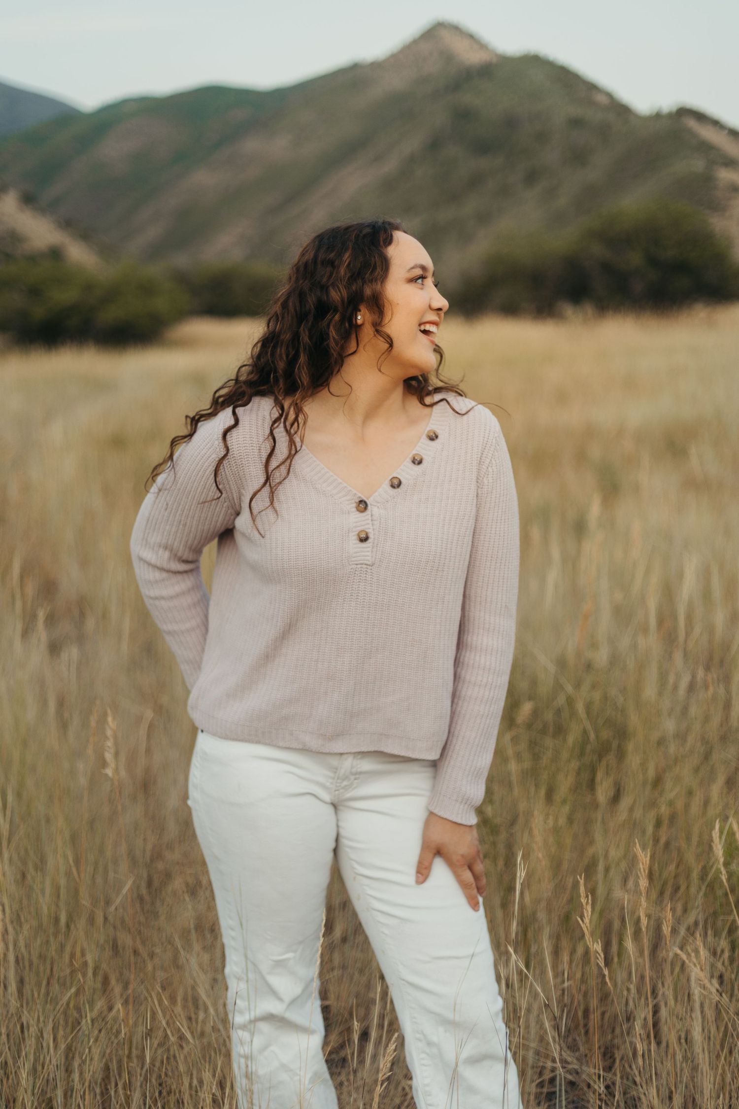 Woman in beige button sweater and white jeans laughing in dry grass field with green mountains.