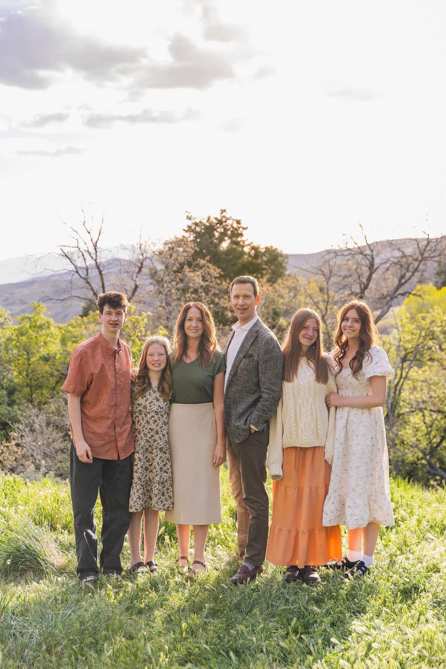 Family of six poses together outdoors in a green hillside setting at golden hour.
