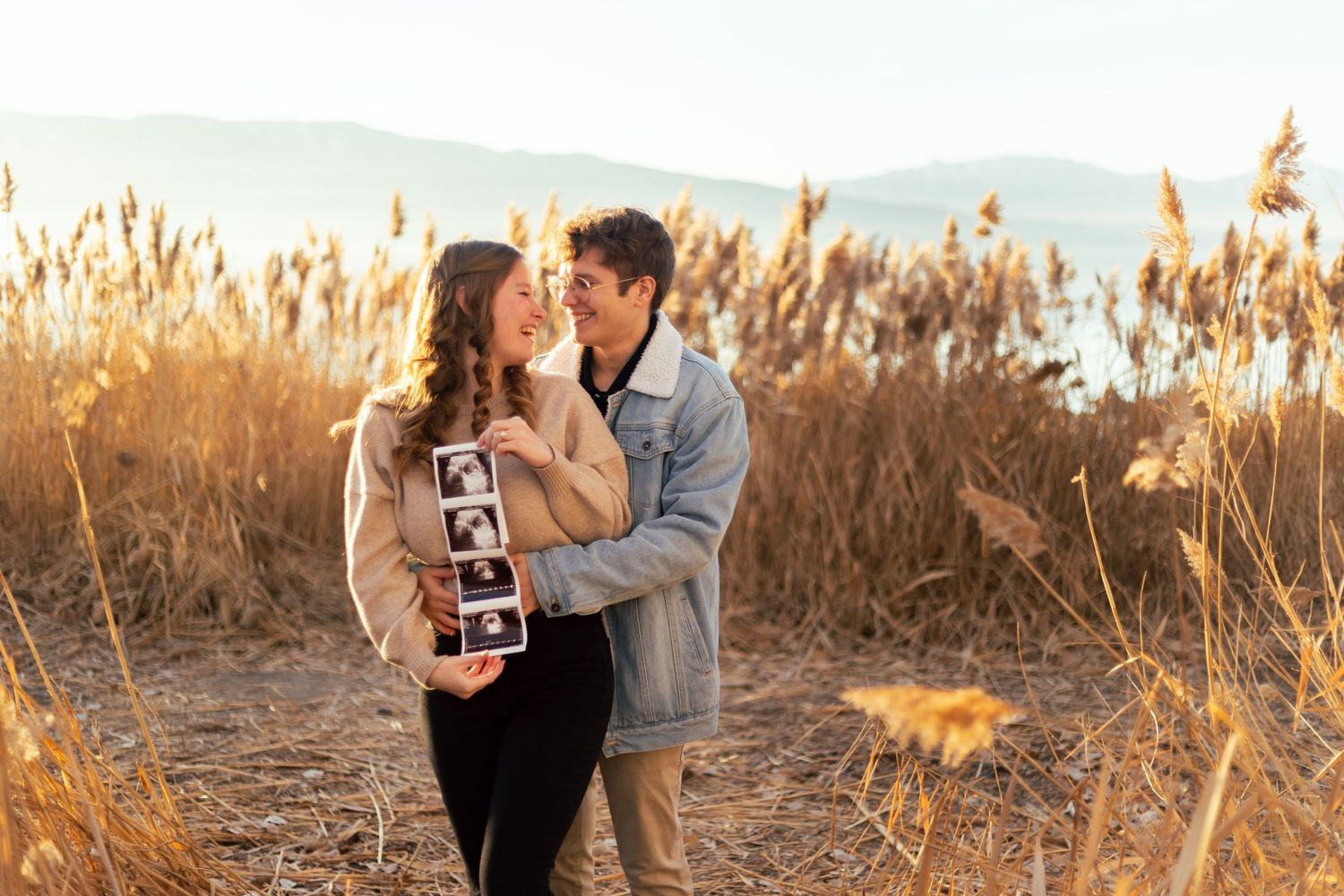 Couple holds ultrasound photos while embracing in a scenic golden field with mountains in the background.