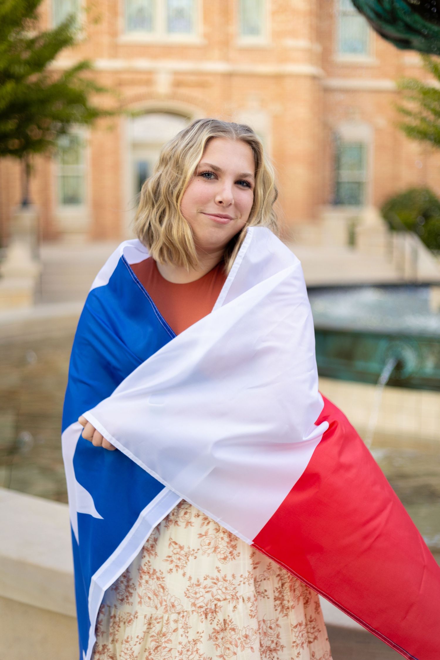 Young blonde woman wrapped in a Texas state flag standing near a fountain outside a brick building.