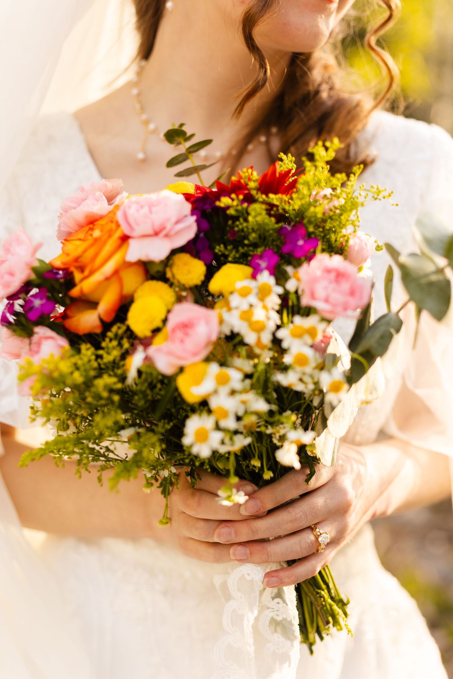 Bride in white lace dress holding vibrant wildflower bouquet with roses, daisies, and colorful blooms.
