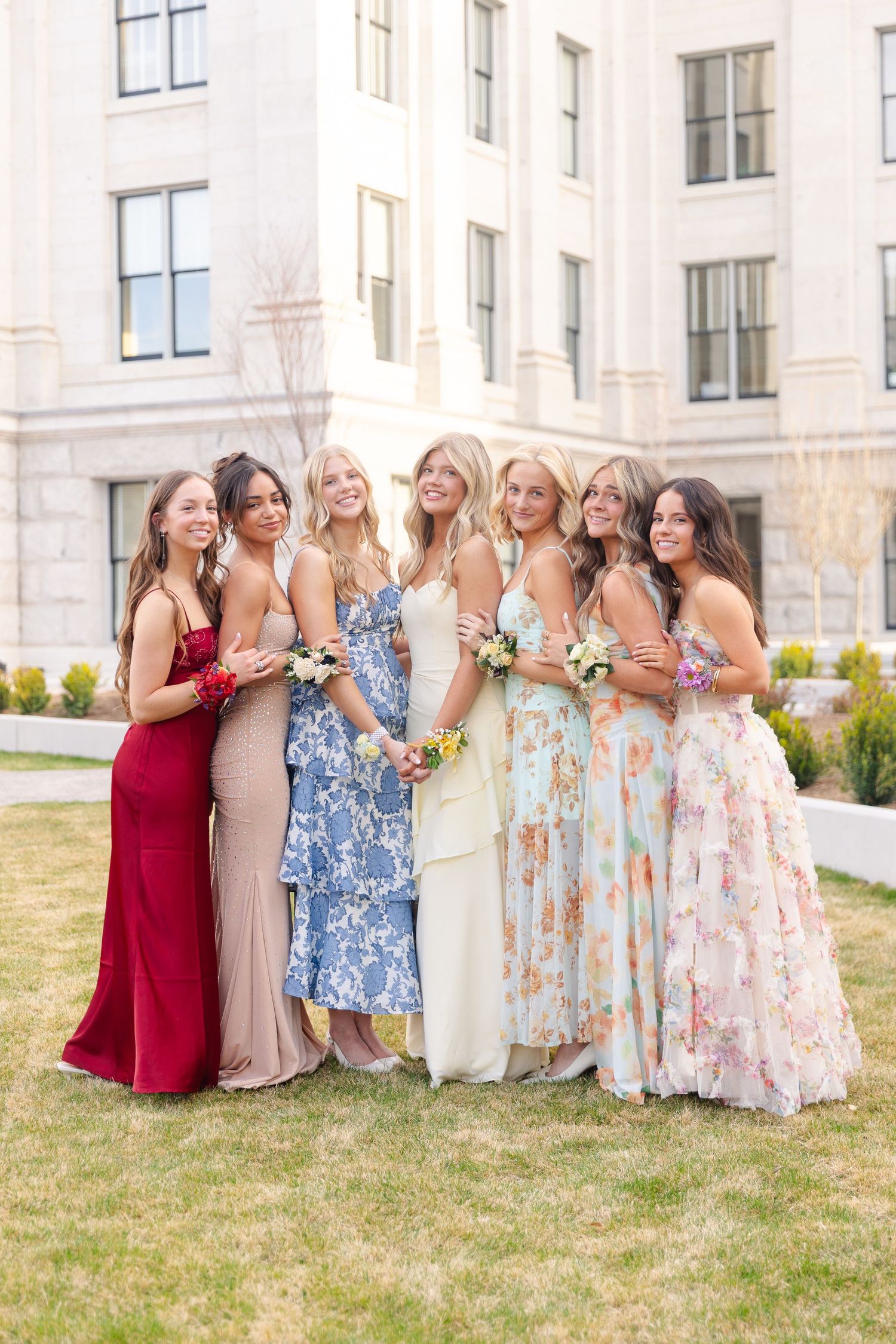 Group of six smiling young women in vibrant formal prom dresses posing outside a building.