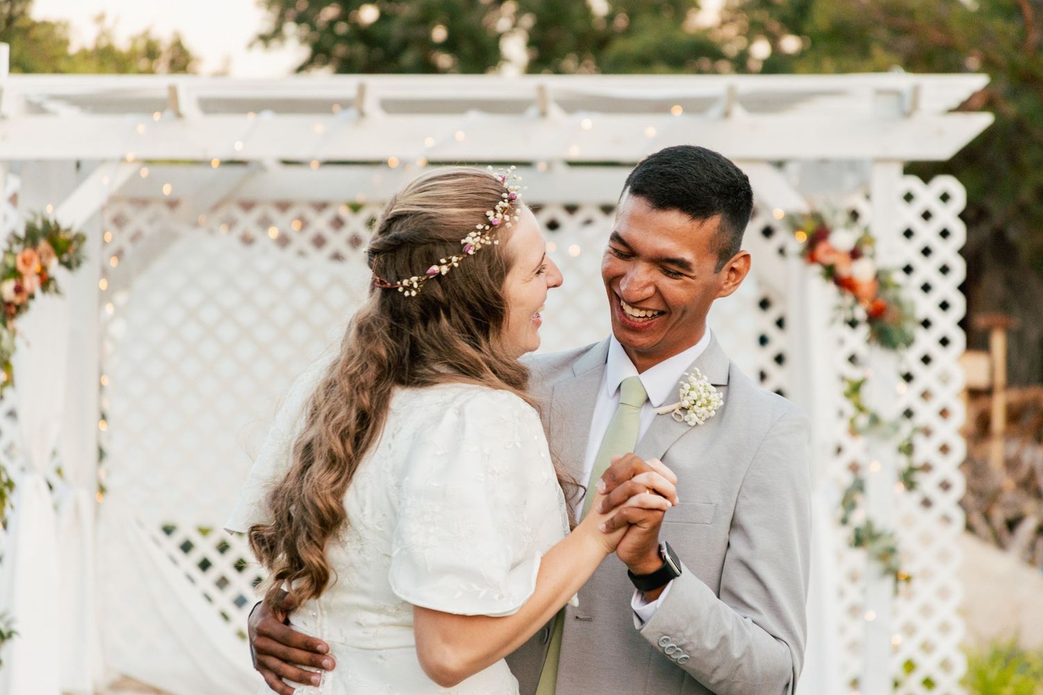 Bride and groom sharing a first dance and smiling at each other under a white garden pergola.