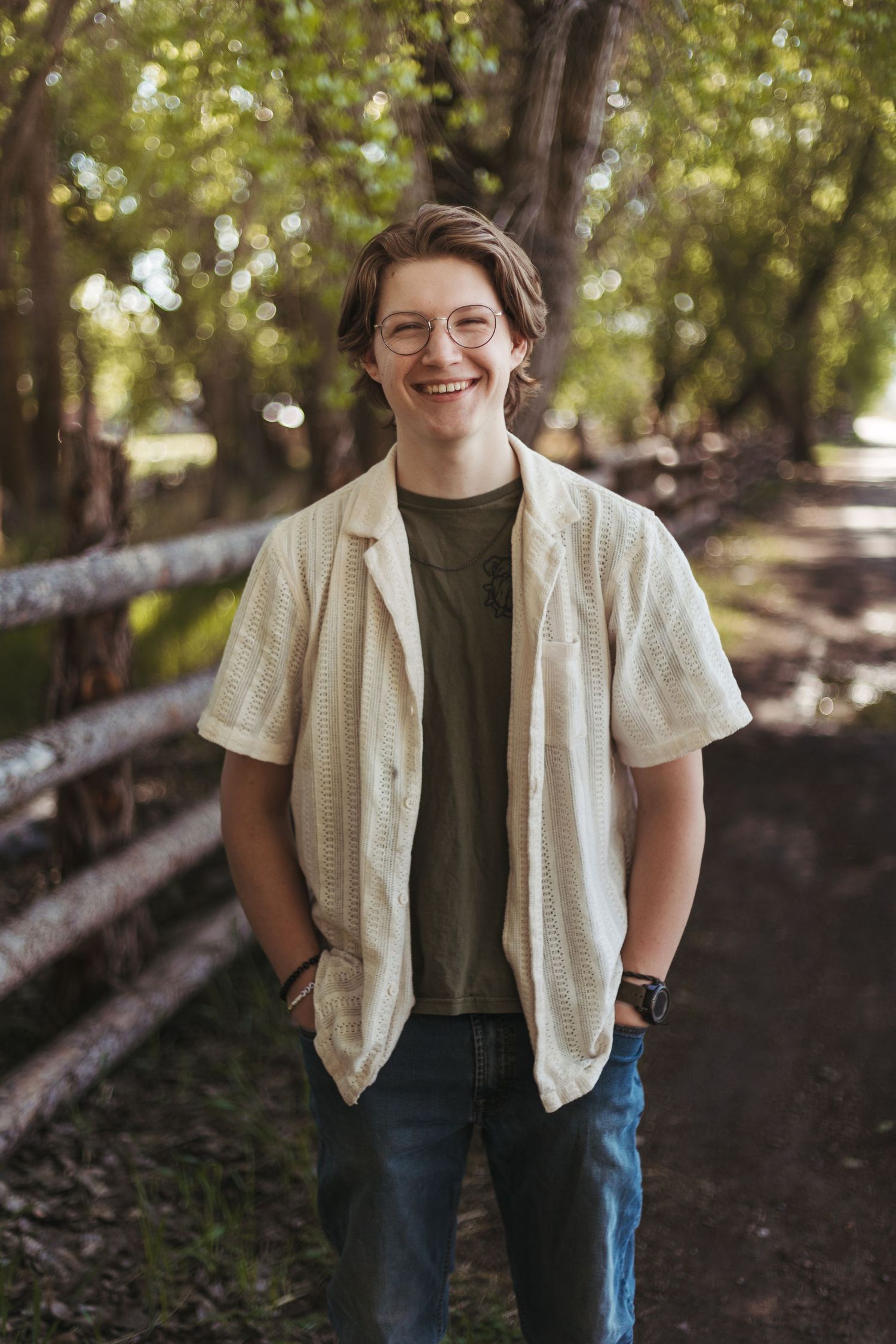 Smiling teenage boy in glasses and casual button-up shirt standing on a tree-lined path.