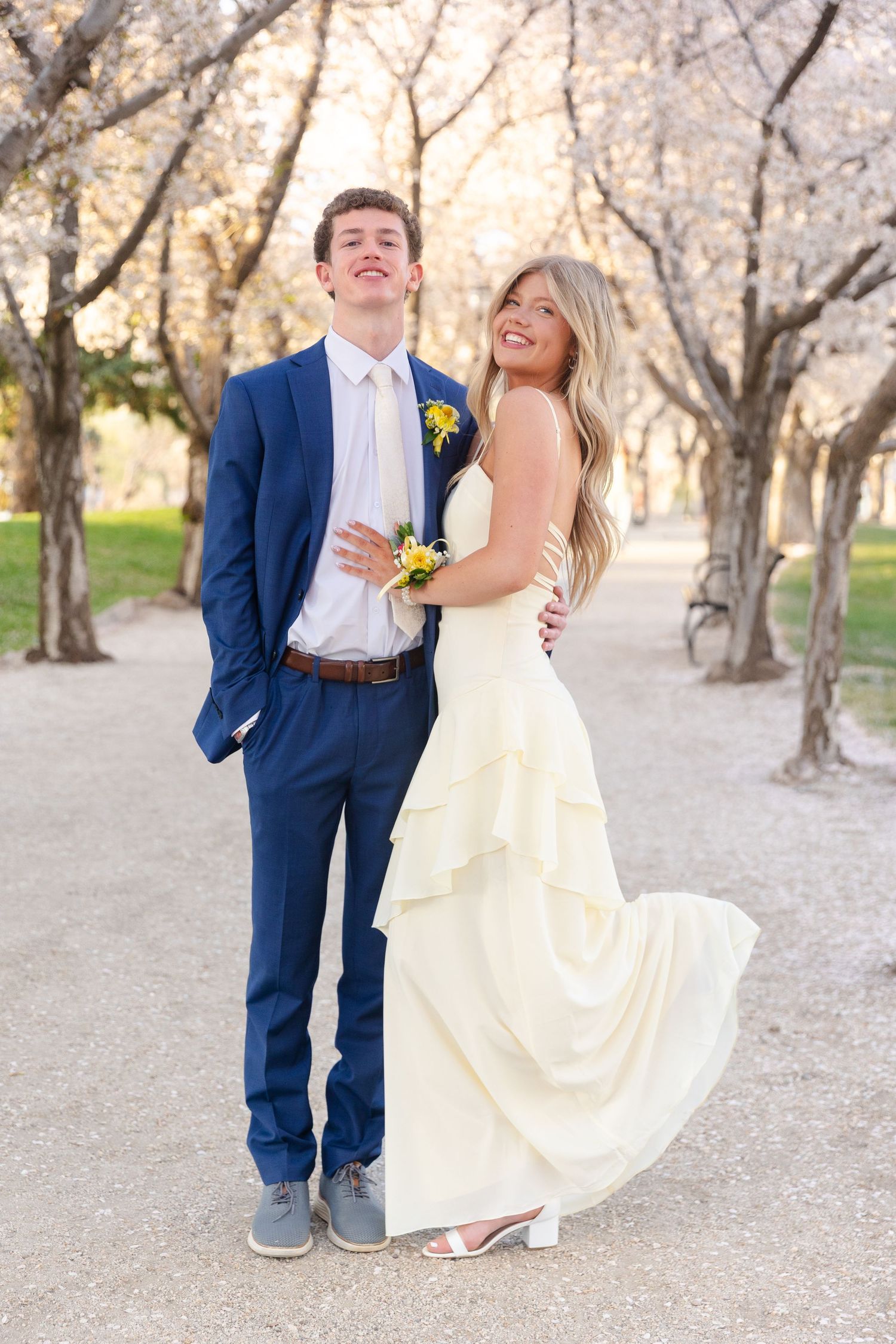 Prom couple posing outdoors among blooming cherry trees, man in navy suit and woman in white gown.