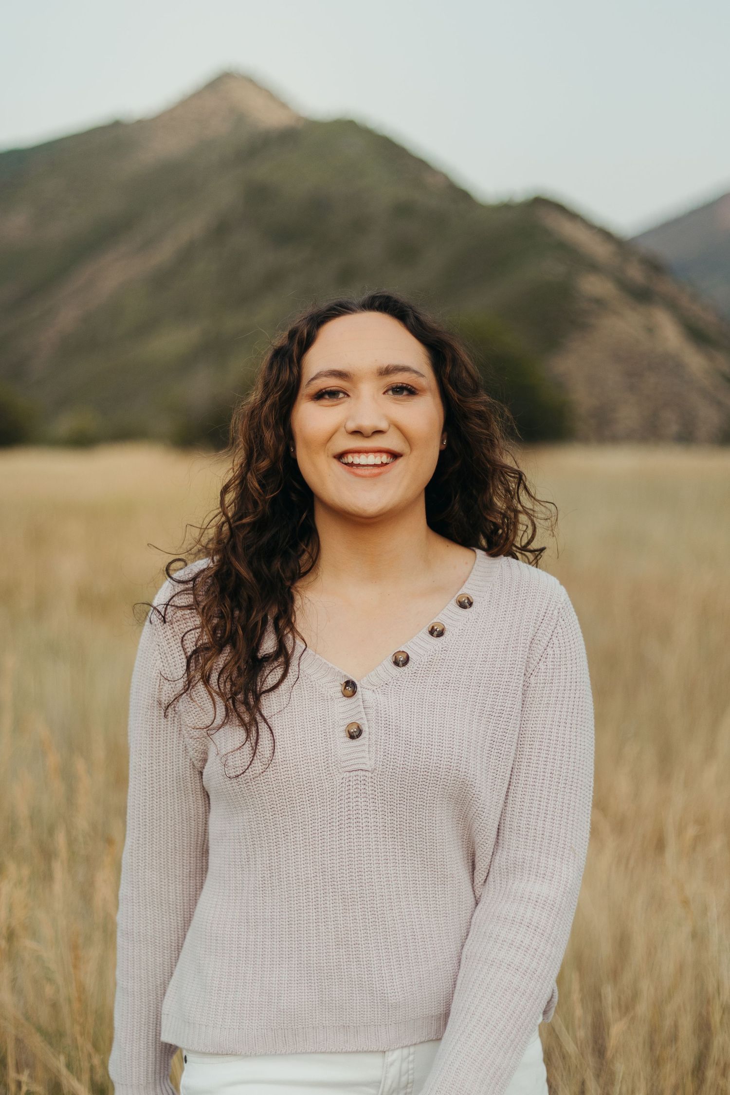 Young woman with curly hair in beige ribbed sweater smiling in golden field with mountain backdrop.