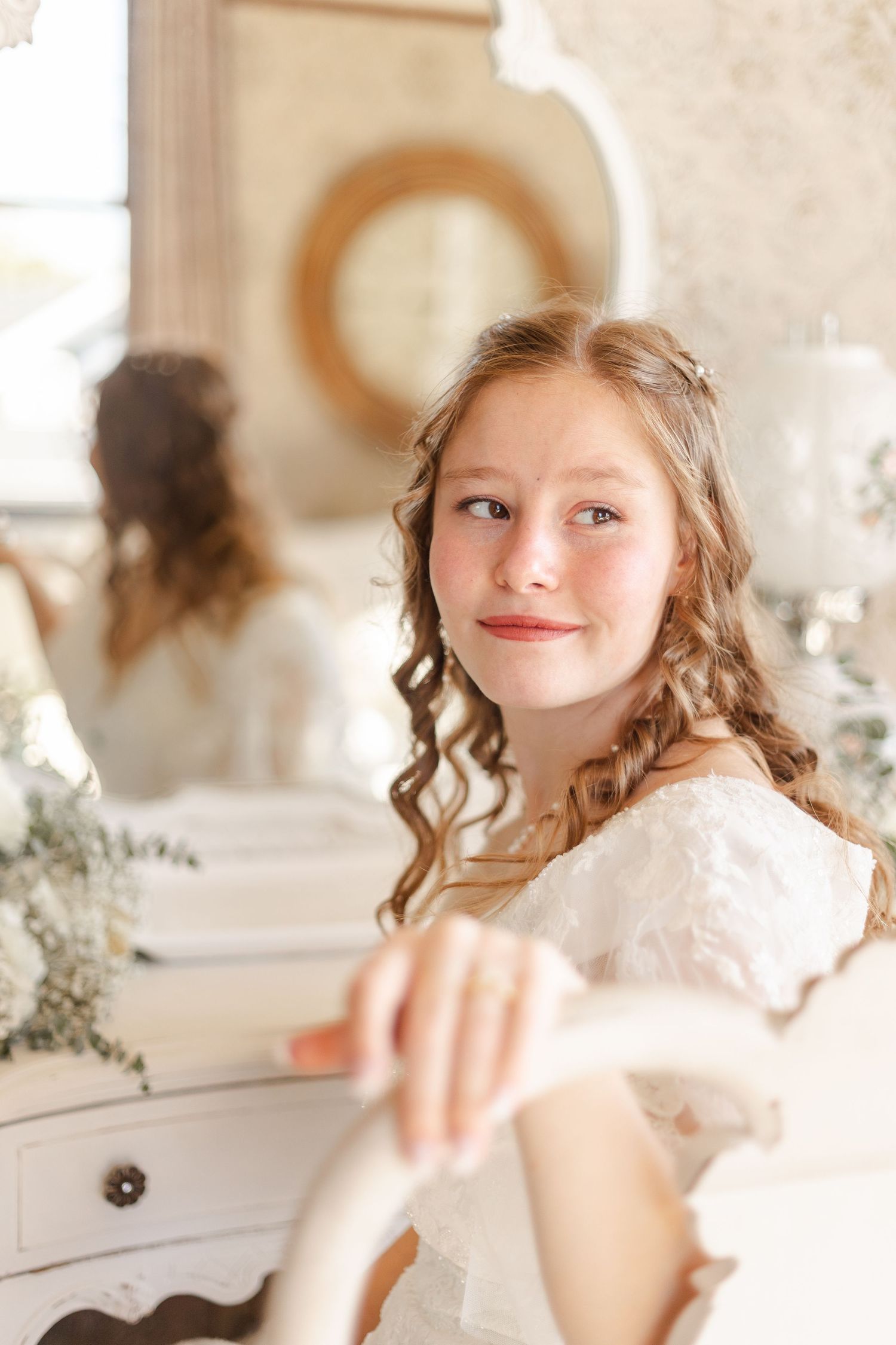 Young bride with curly hair smiles softly while sitting near a vanity mirror in a bright room.