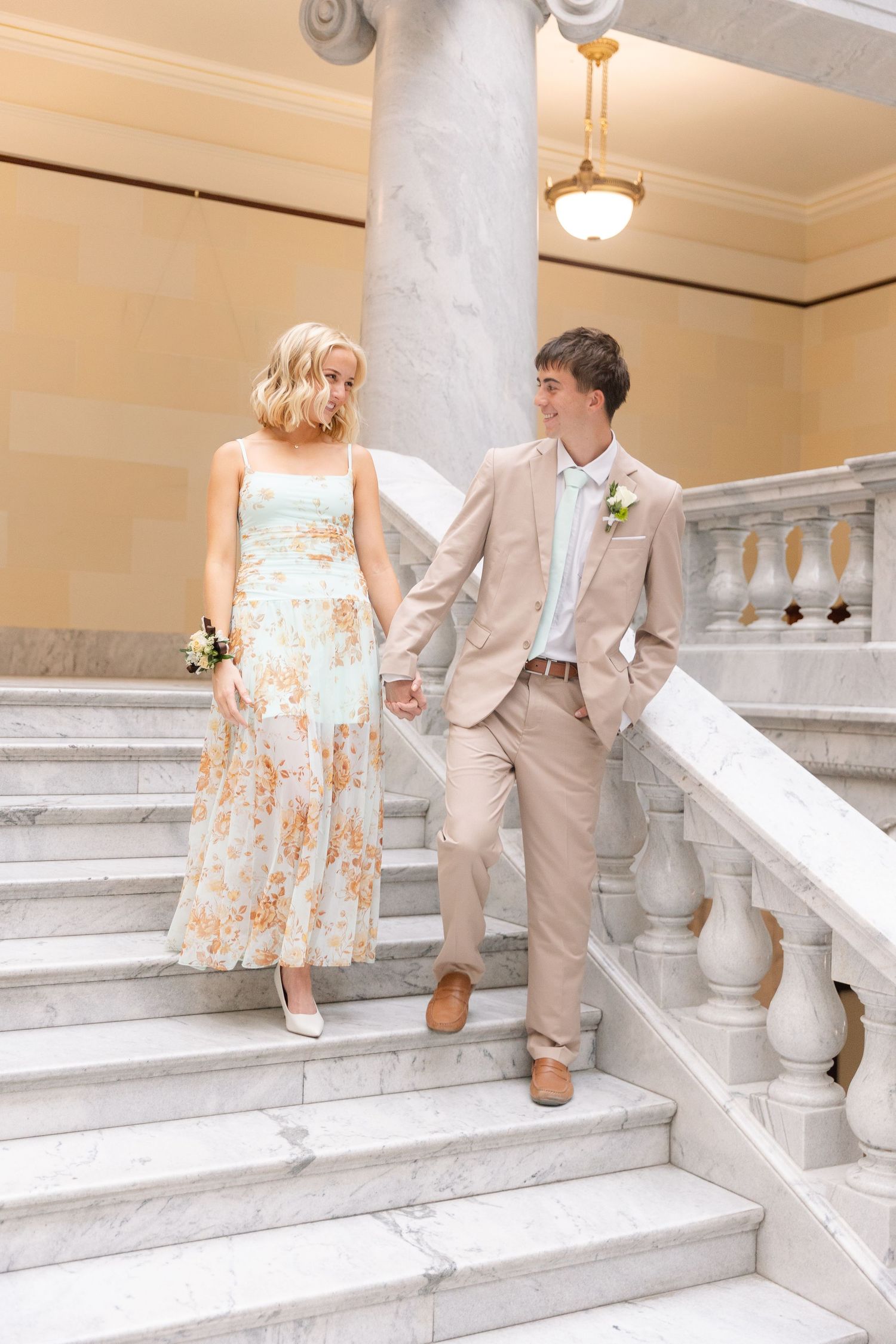 Couple in formal attire holding hands on marble staircase, woman in floral dress and man in tan suit.