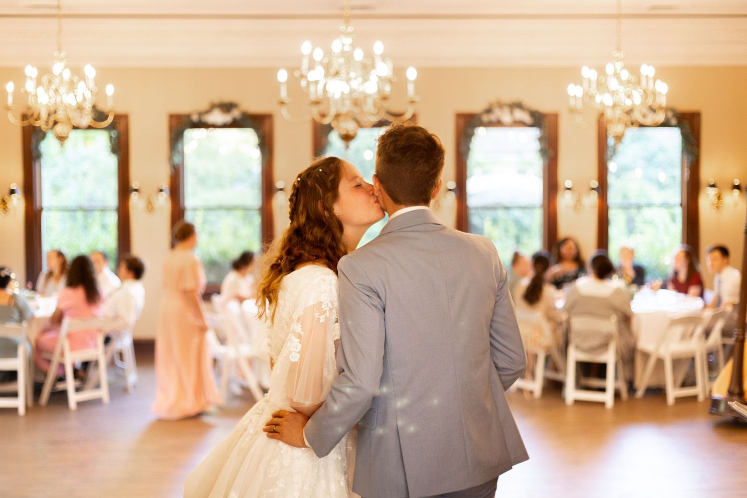Bride and groom kissing during first dance in grand ballroom with chandeliers and seated wedding guests.