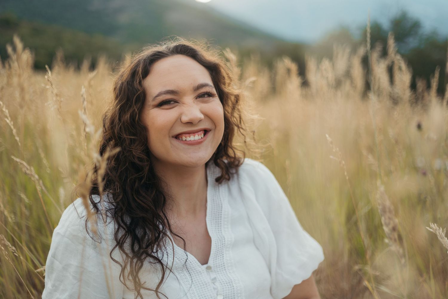 Smiling young woman with curly hair in white top sitting in golden field with mountains behind.