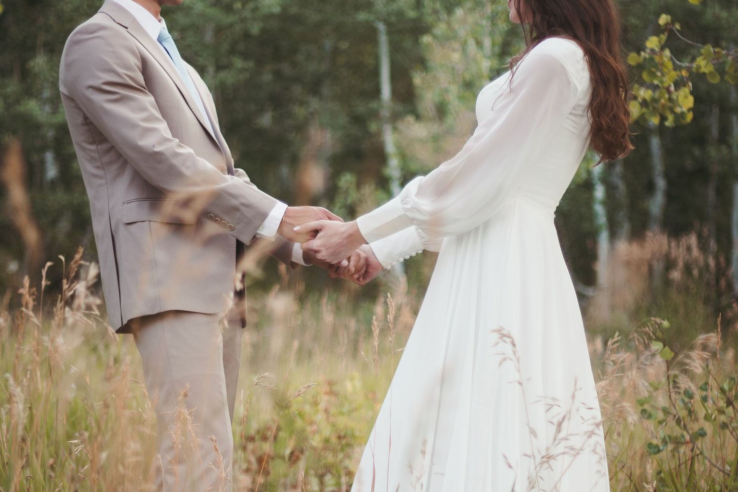 Bride and groom holding hands walking through a sunlit field of tall golden grass.