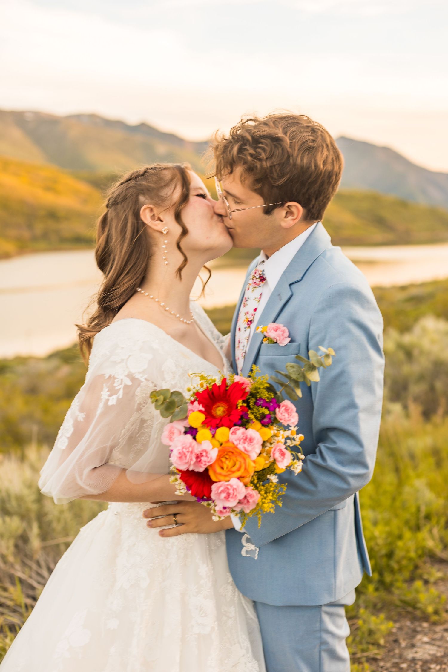 Bride and groom kissing with colorful bouquet against scenic mountain lake backdrop at sunset.