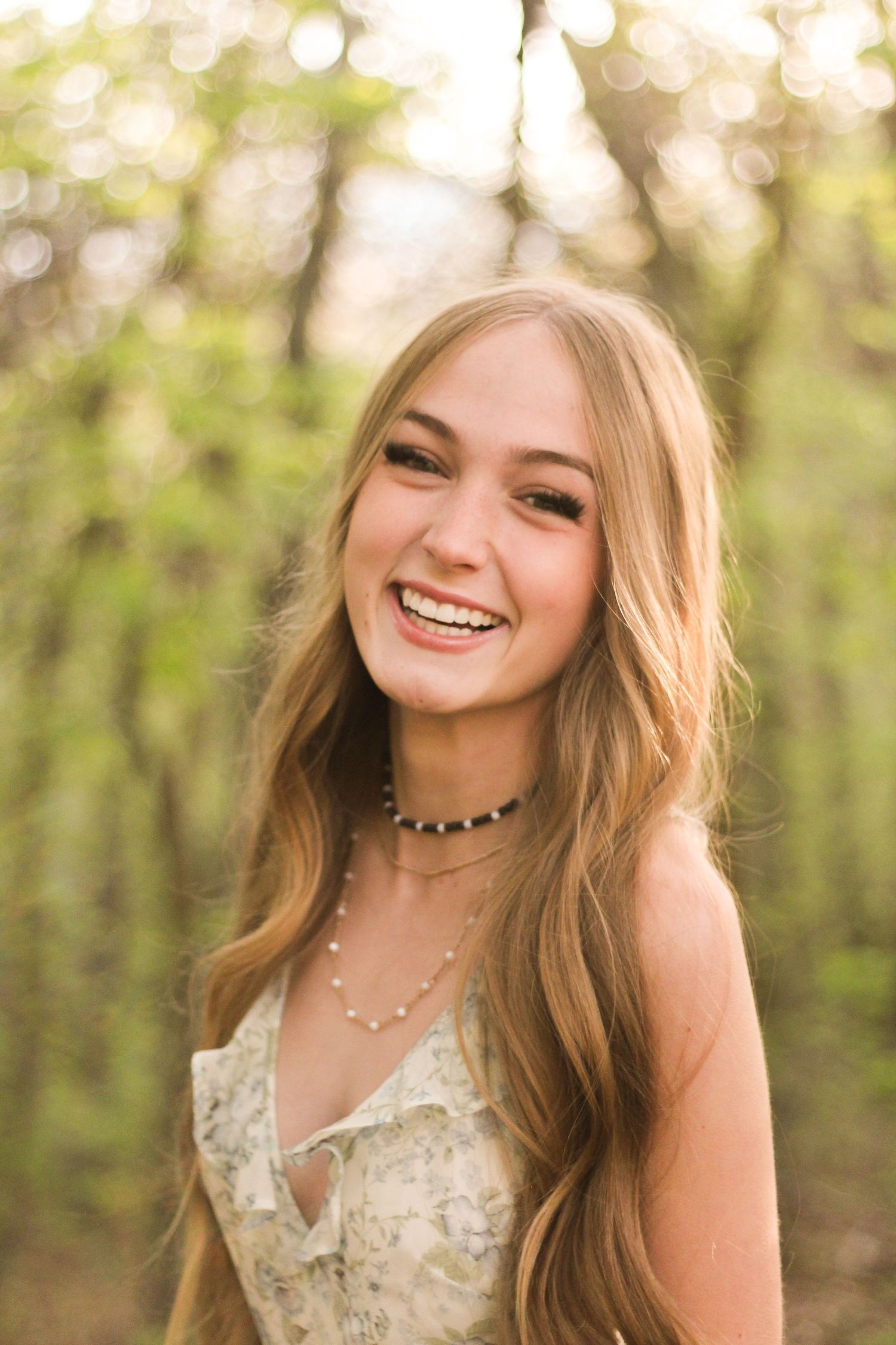 Smiling young woman with long blonde hair wearing floral dress and layered necklaces in sunlit forest.