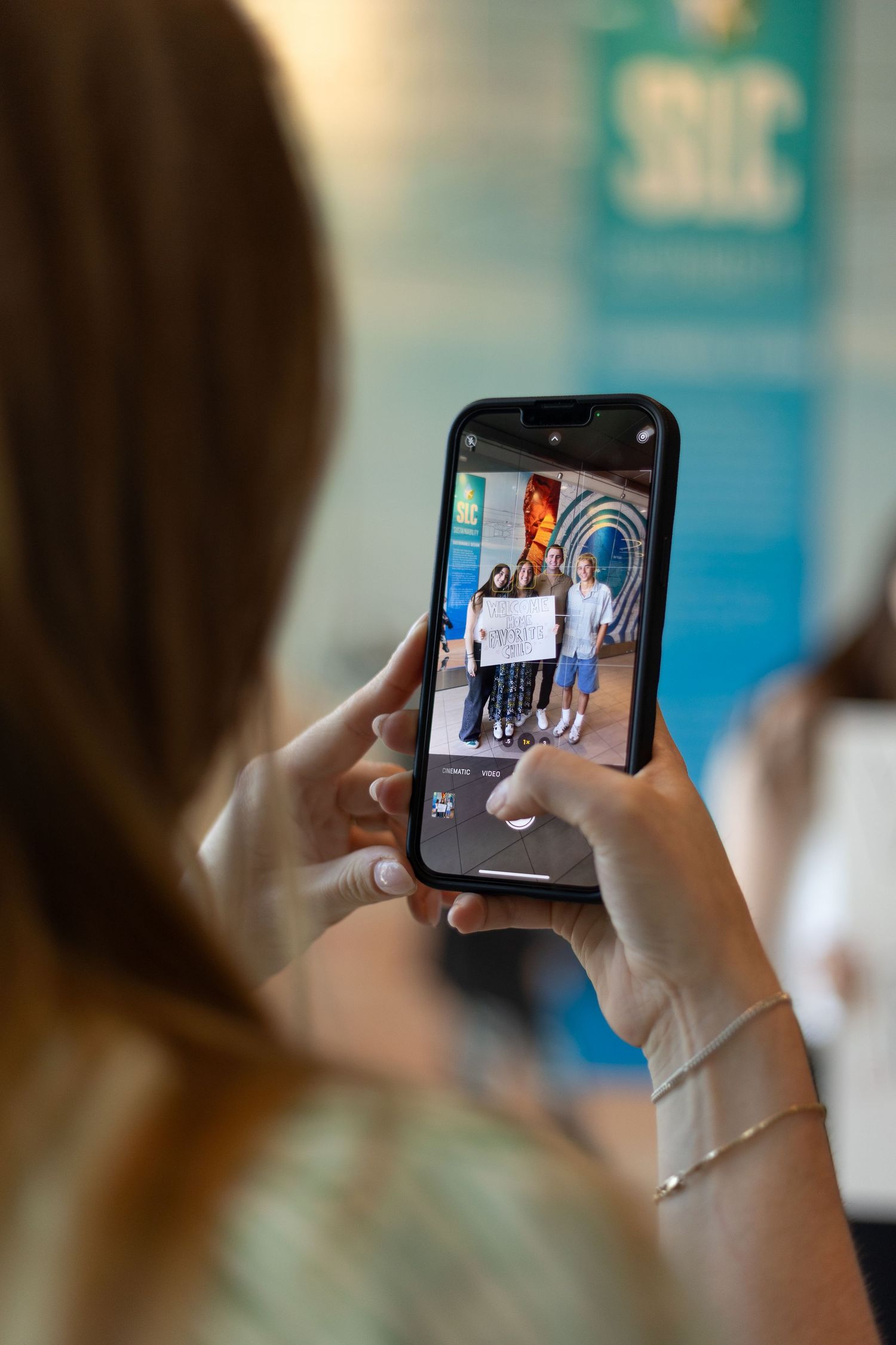 Woman photographing a group holding welcome home signs on a smartphone at an indoor event.
