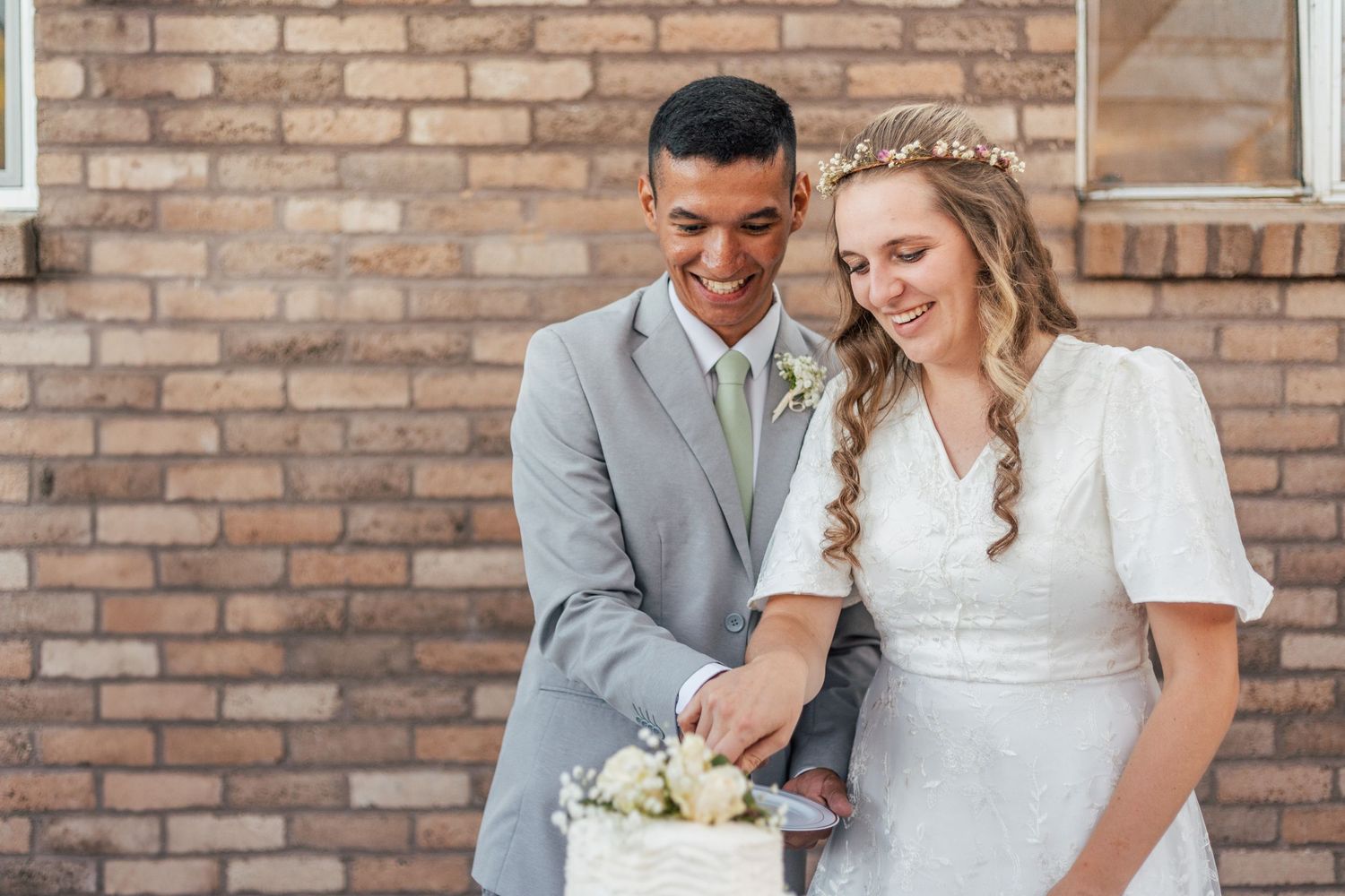 Bride and groom smiling while cutting their white wedding cake together against a brick wall.