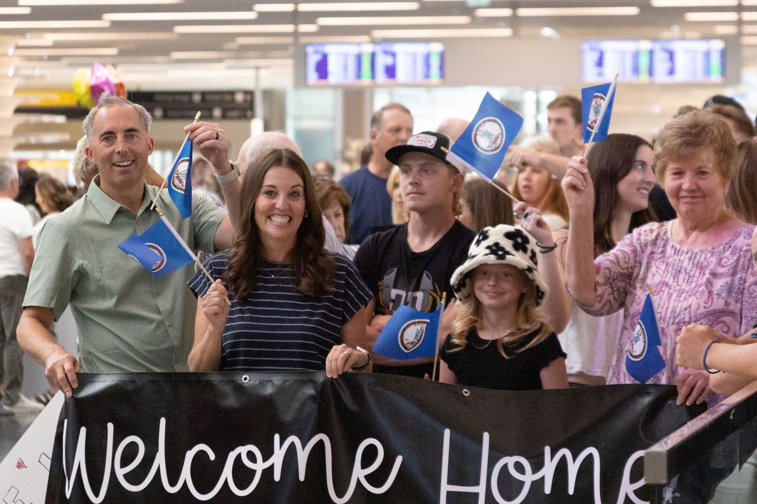 Excited family holds a Welcome Home banner and blue flags greeting a returning missionary at the airport.
