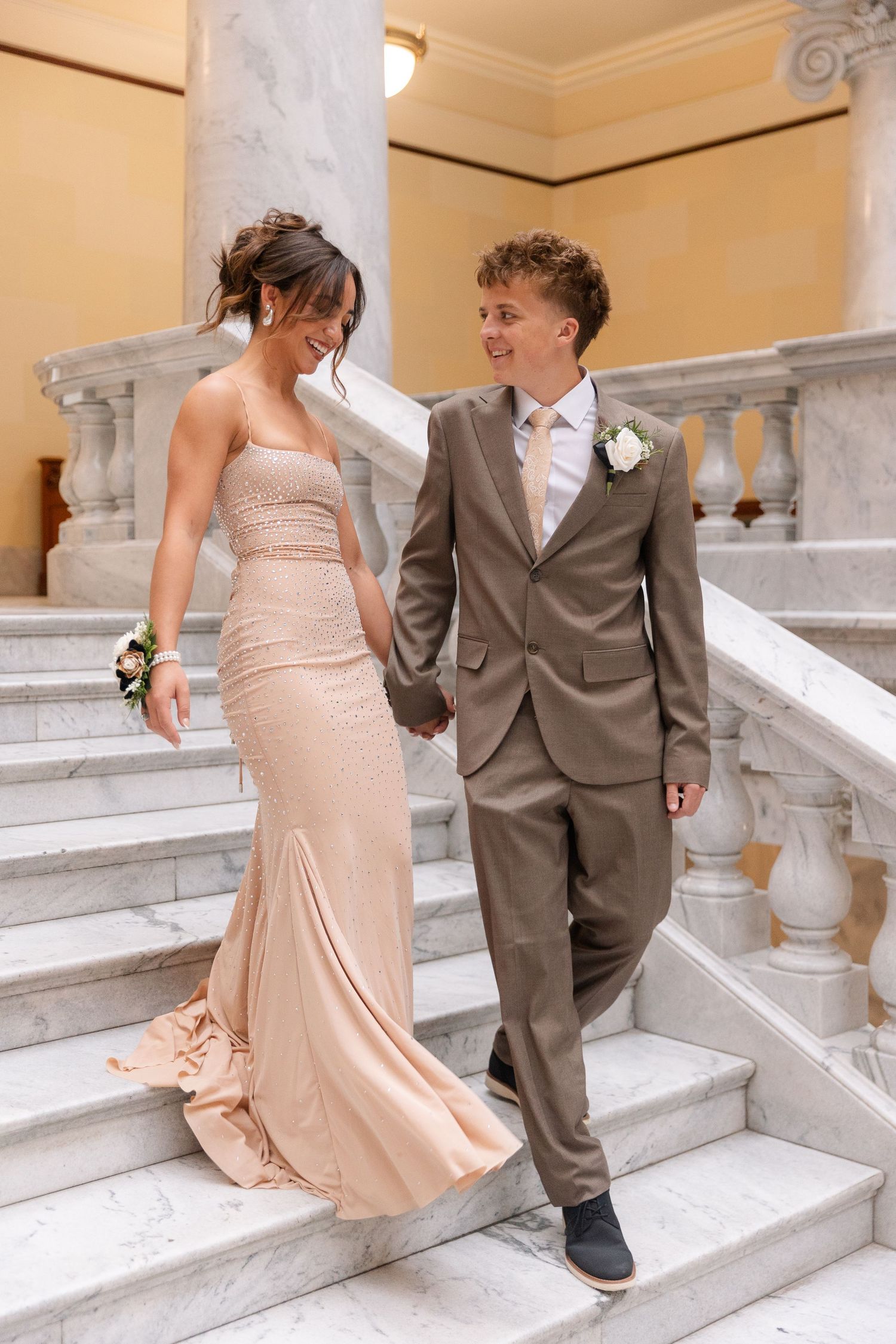 Elegant prom couple in formal wear holding hands on marble staircase, woman in blush gown and man in tan suit with boutonniere.