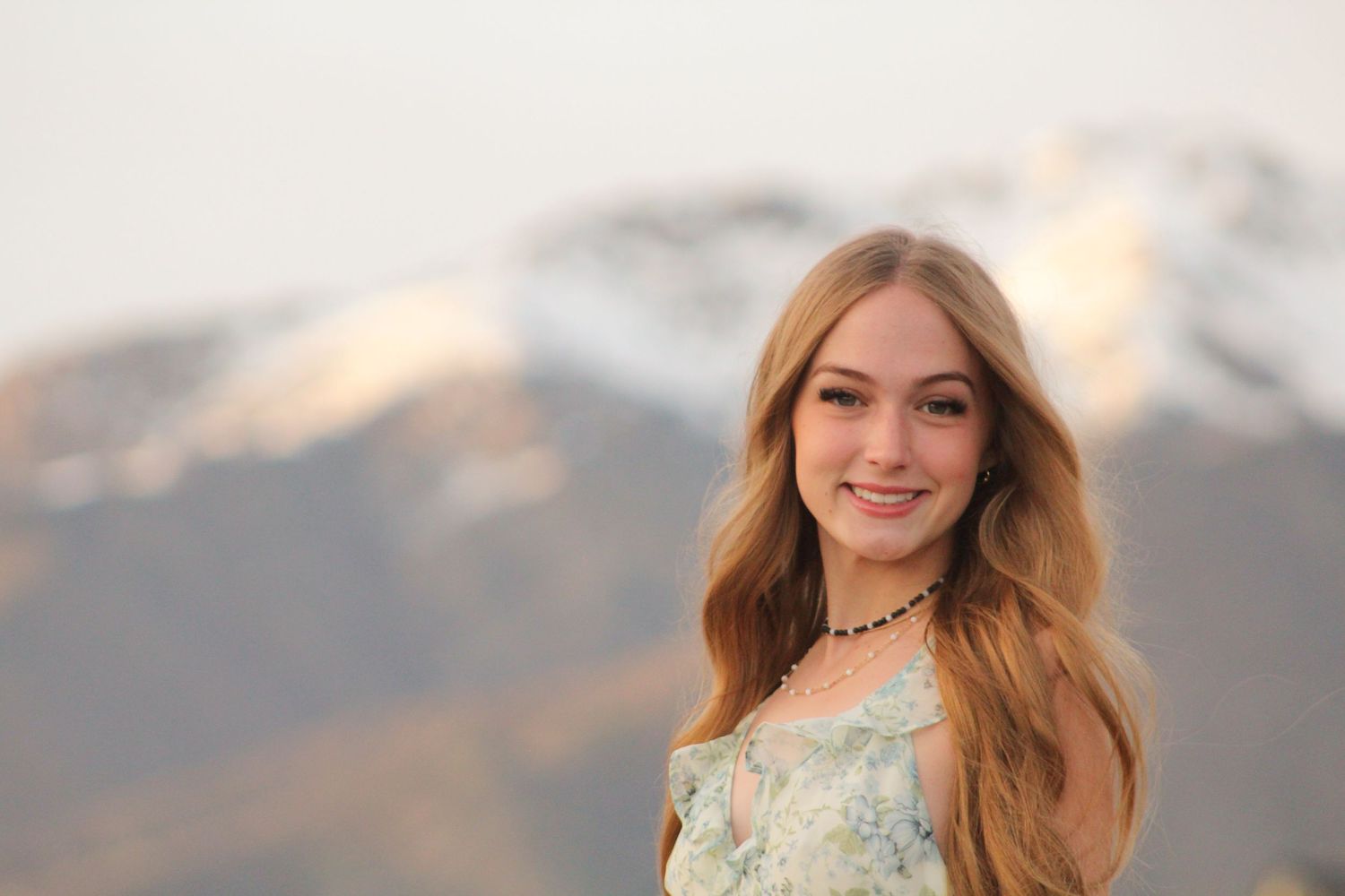 Young woman with long blonde hair smiling outdoors with snow-capped mountains in the background.