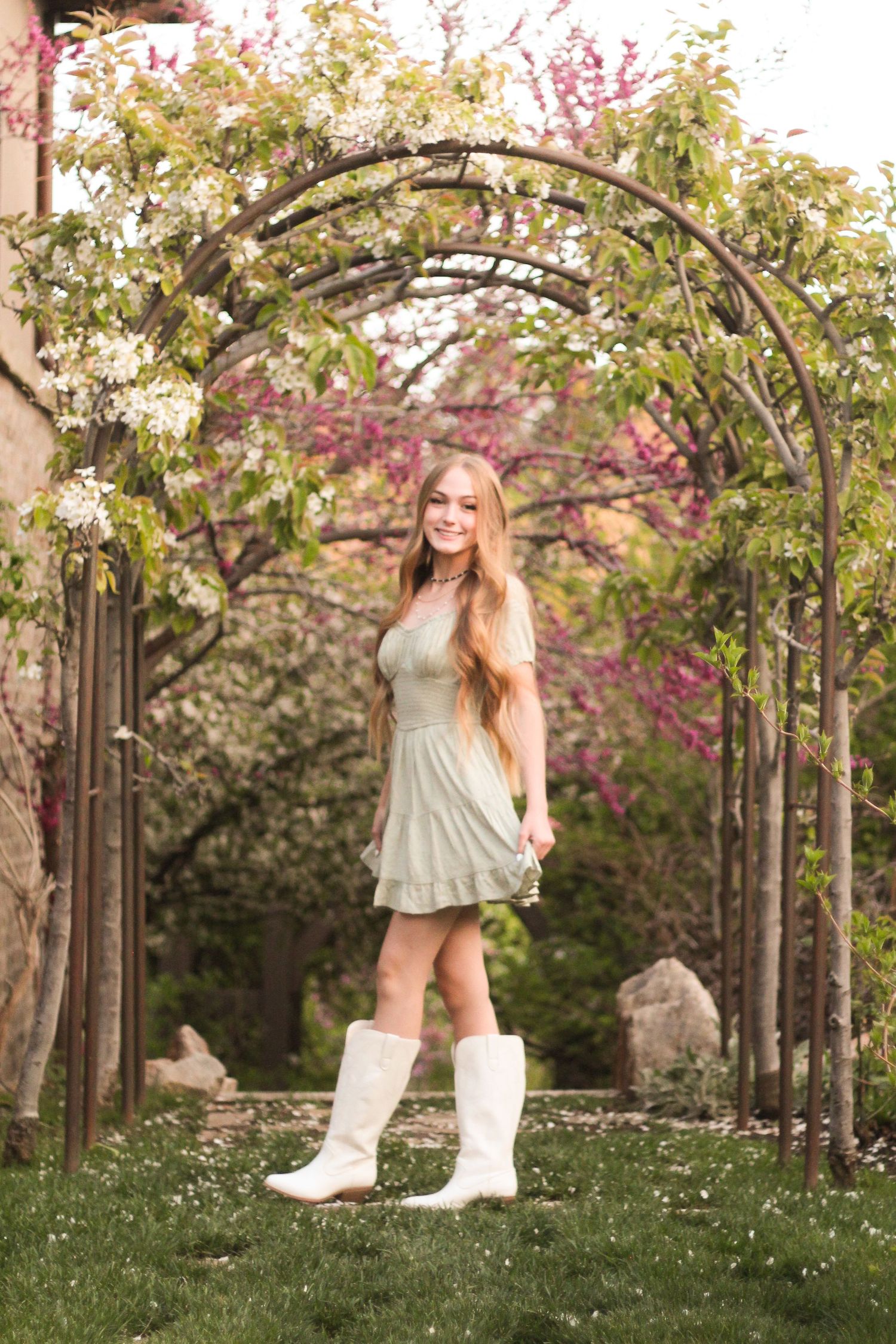 Young woman in a sage green dress and white cowboy boots standing under a floral garden archway in spring.