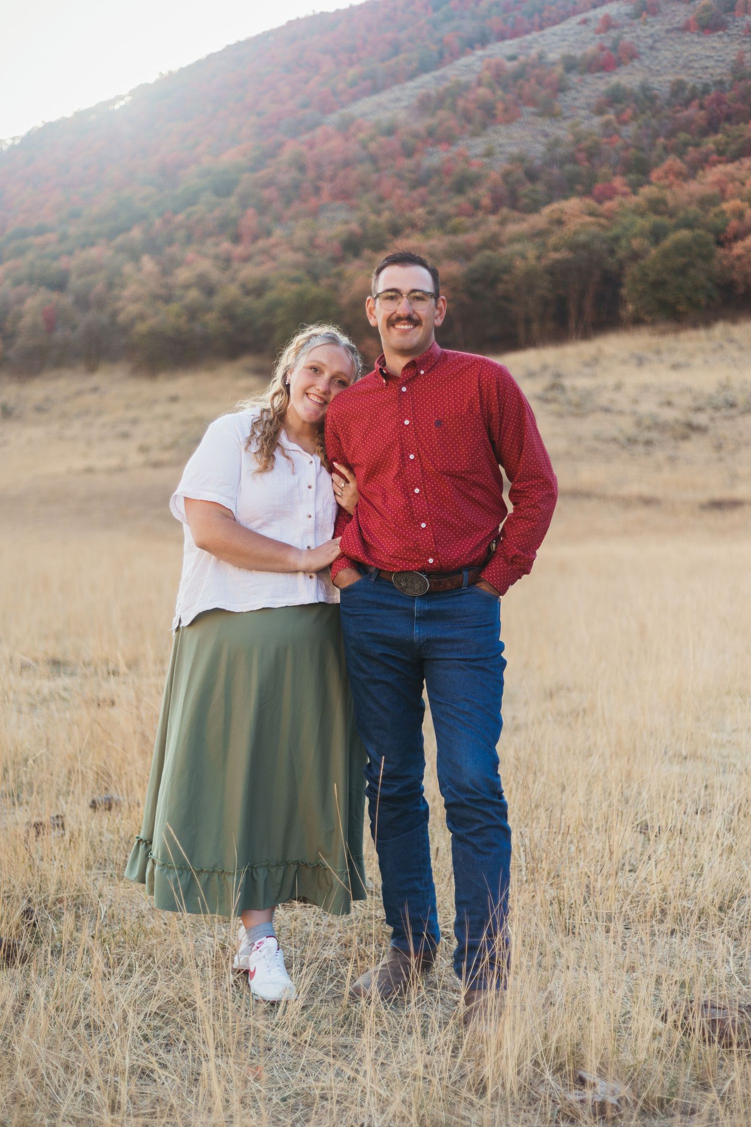 Couple smiling together in a golden field with autumn foliage on a hillside behind them.