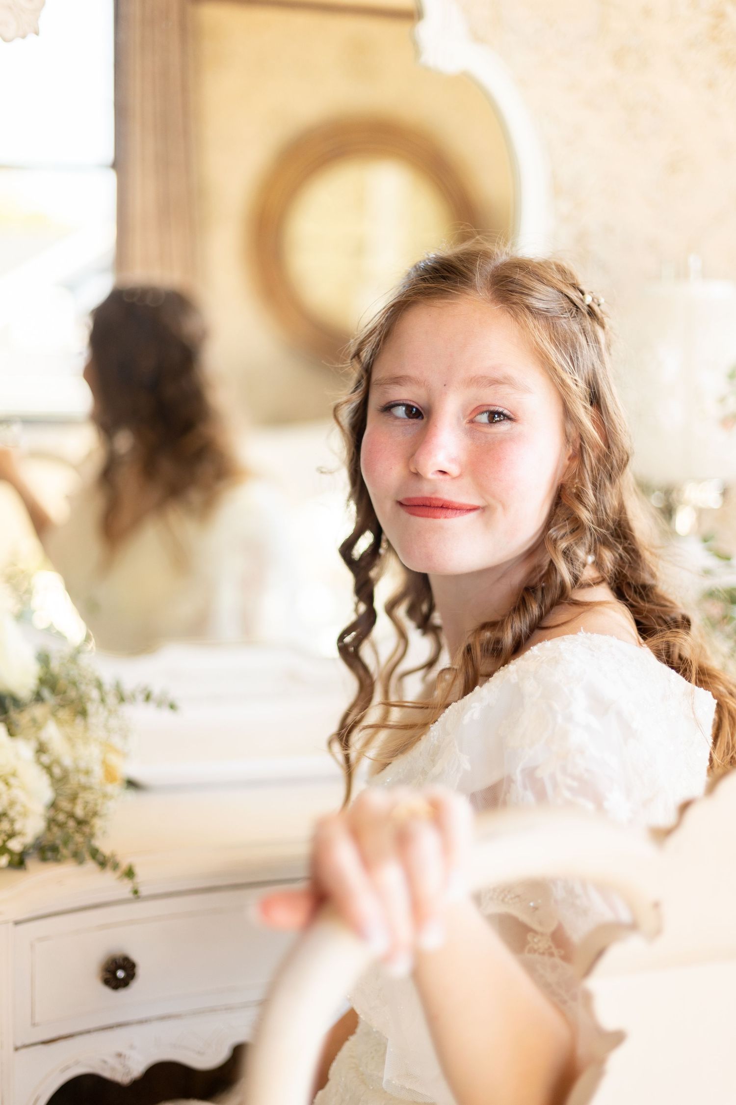 Young girl with curly hair in a white dress smiling softly while seated near a vintage mirror.