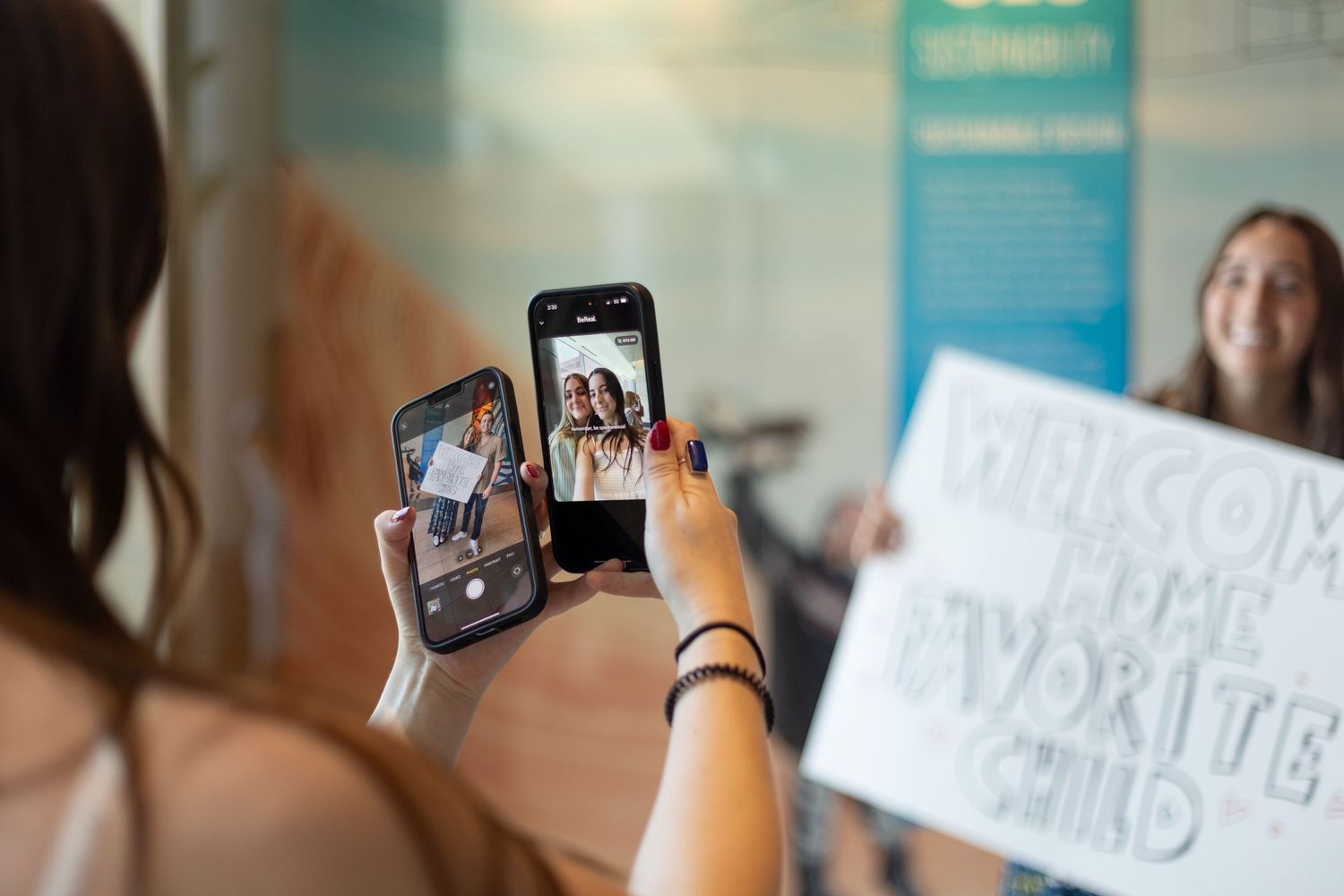 Person photographing a smiling woman holding a welcome home sign using two smartphones.