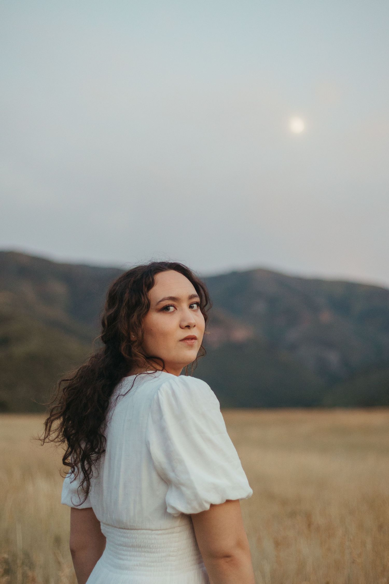 Young woman in white puff-sleeve dress looking back over shoulder in golden field with full moon.