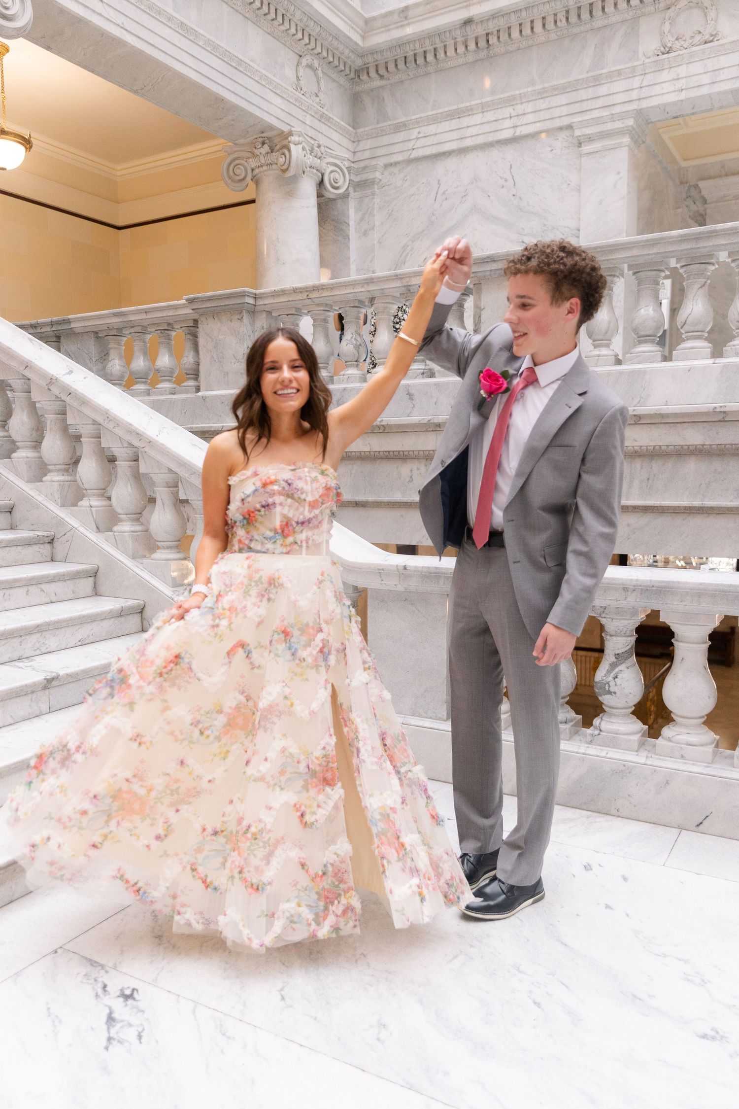 Prom couple dancing on marble staircase, girl in colorful floral ballgown and boy in gray suit with pink tie at the Utah Capitol.