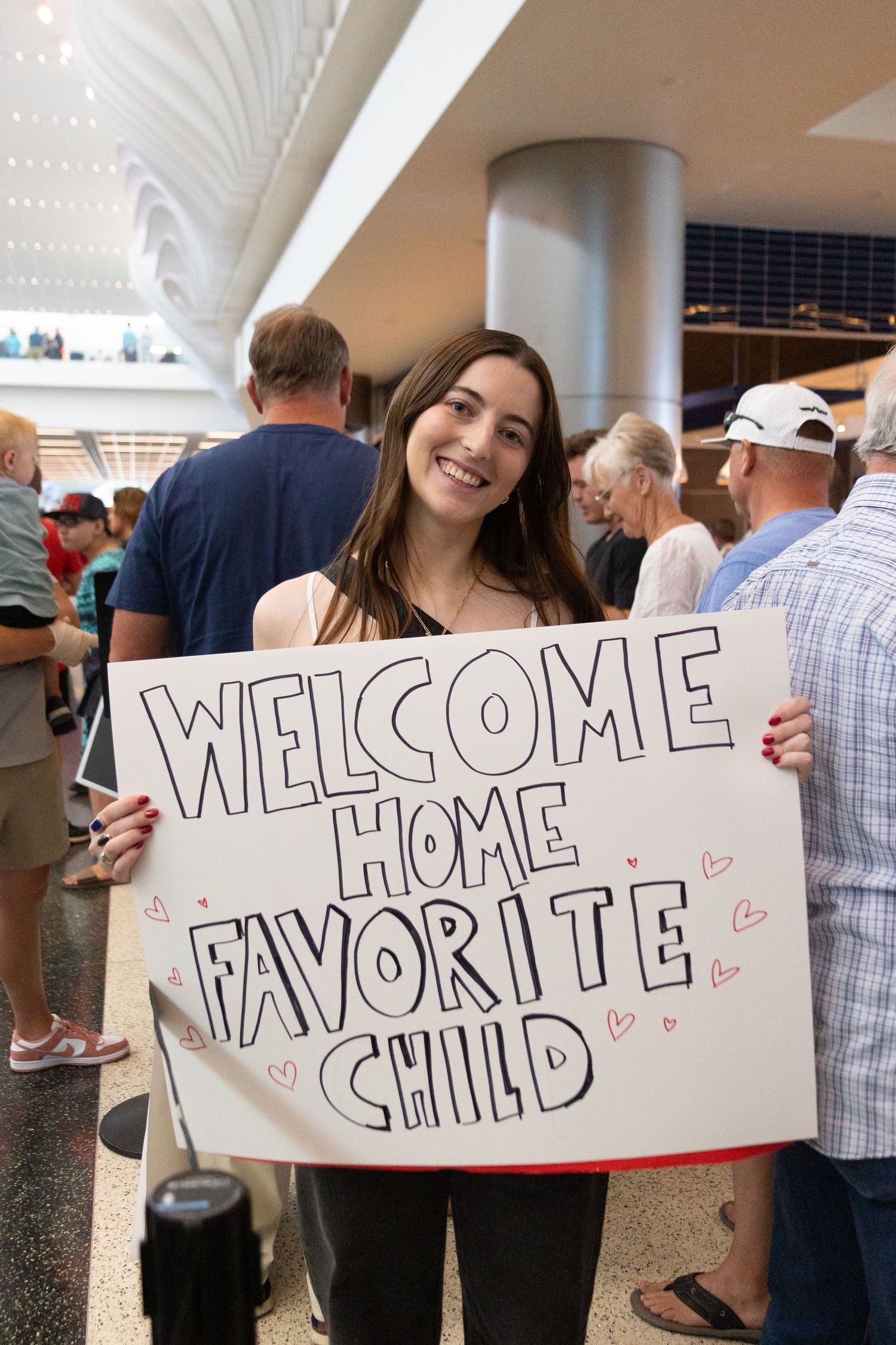 Smiling brunette woman holds a handmade Welcome Home Favorite Child sign at an airport arrivals area for her returned missionary sister.