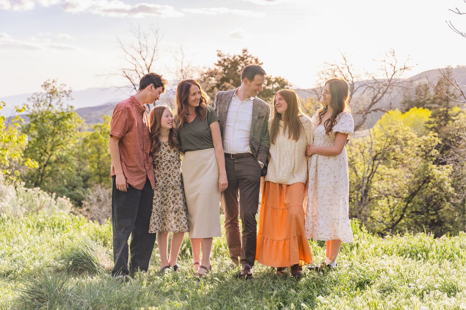 Six family members smiling and embracing outdoors in a sunny meadow with trees and mountains in the background.